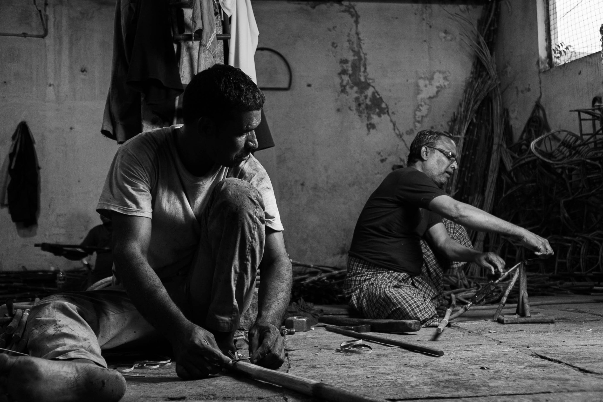 Two men working on a construction or metalwork project in a dimly lit indoor space with a rough wall and various materials around them.