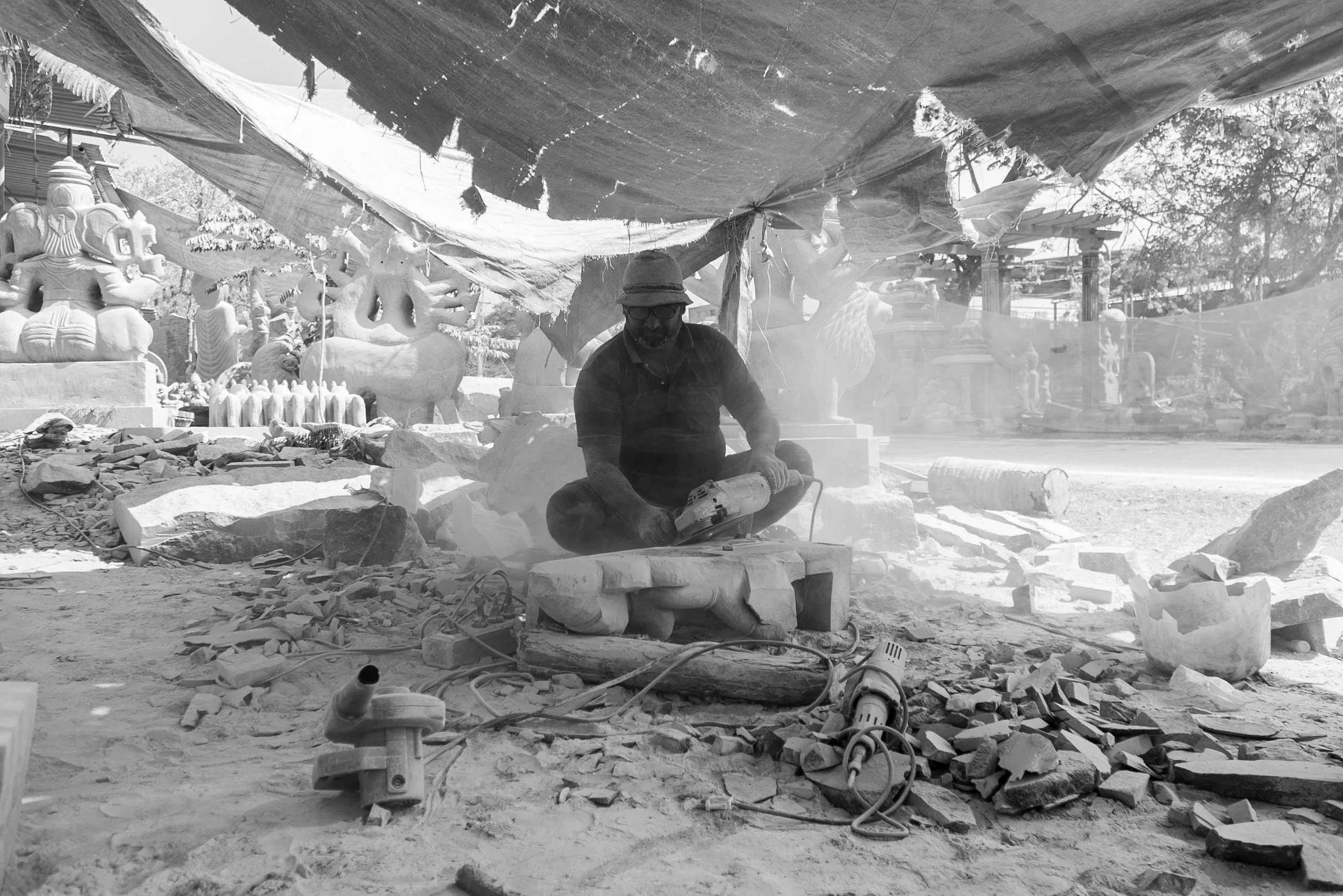 A man working on carving a stone sculpture outdoors under a makeshift canopy, surrounded by stone statues and debris.