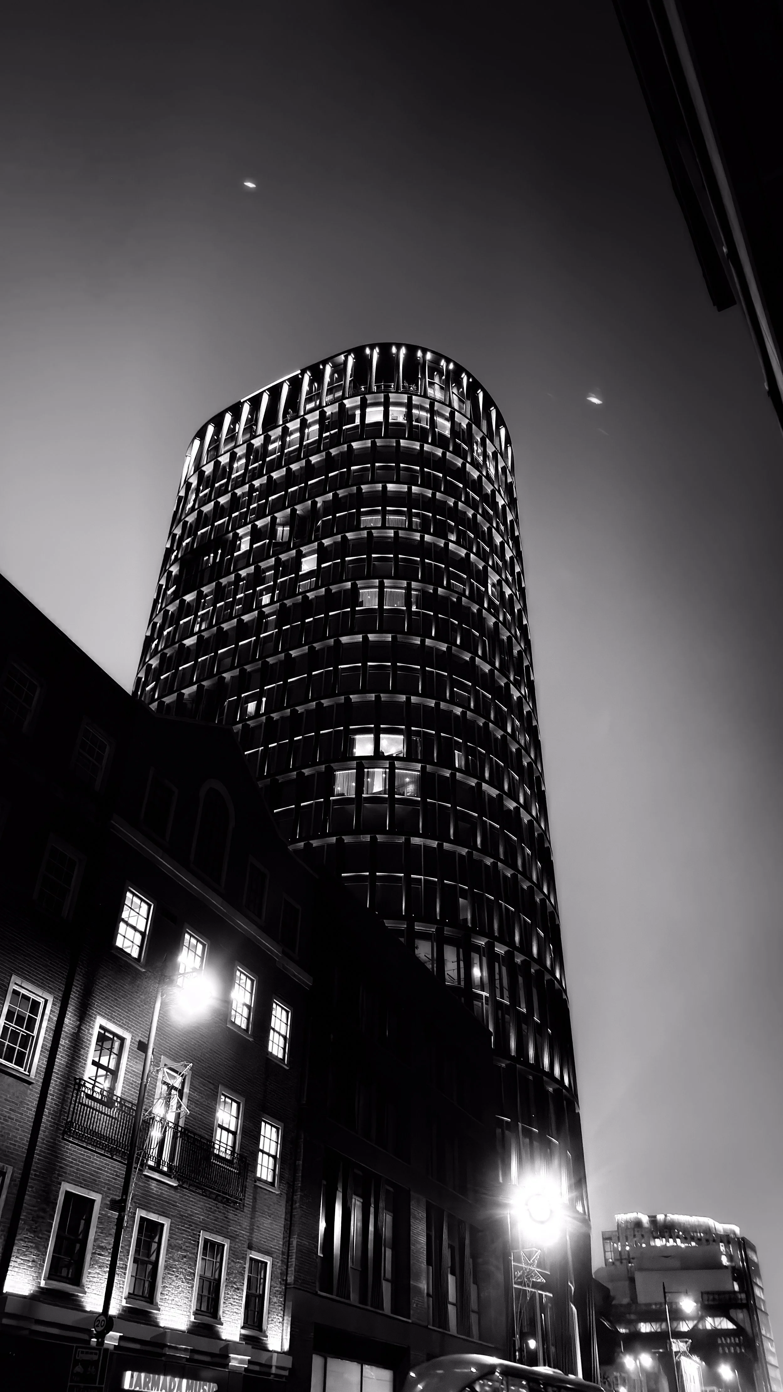 Nighttime cityscape featuring a modern, tall, cylindrical glass skyscraper illuminated from within, alongside shorter traditional brick buildings with lit windows and streetlights.
