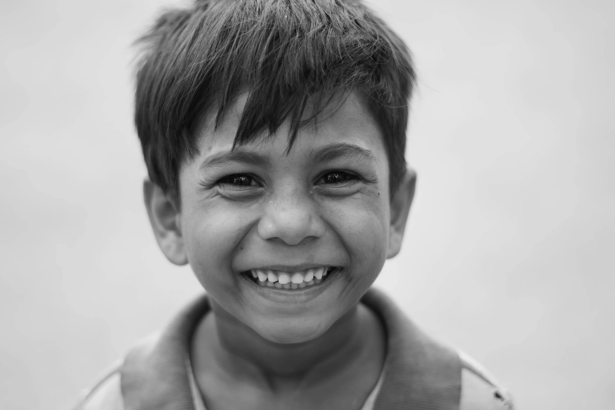 Black and white close-up photo of a smiling young boy with short hair, wearing a shirt and backpack, against a plain background.