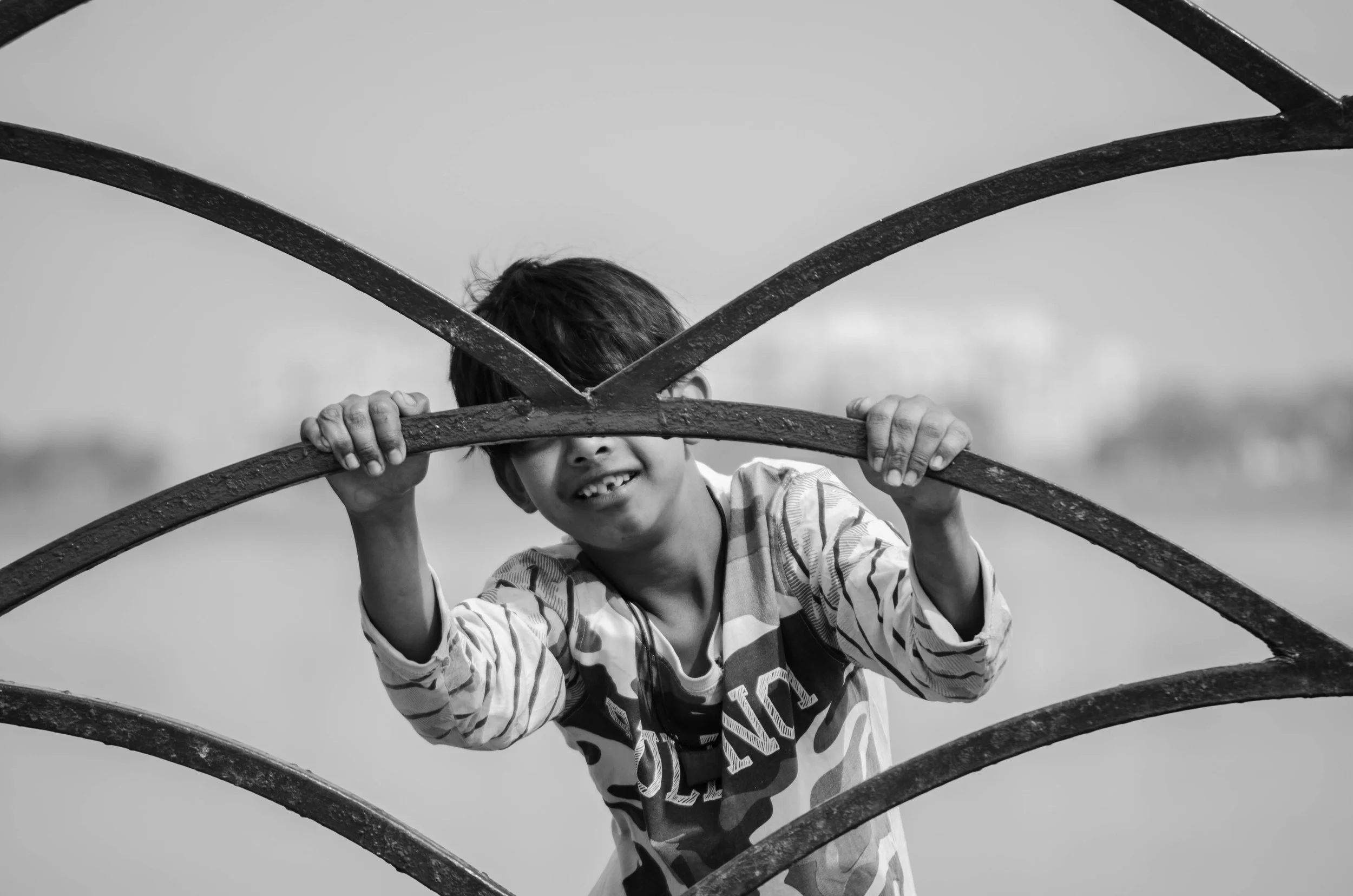 Young boy holding onto a metal fence, smiling, in a black-and-white photo.