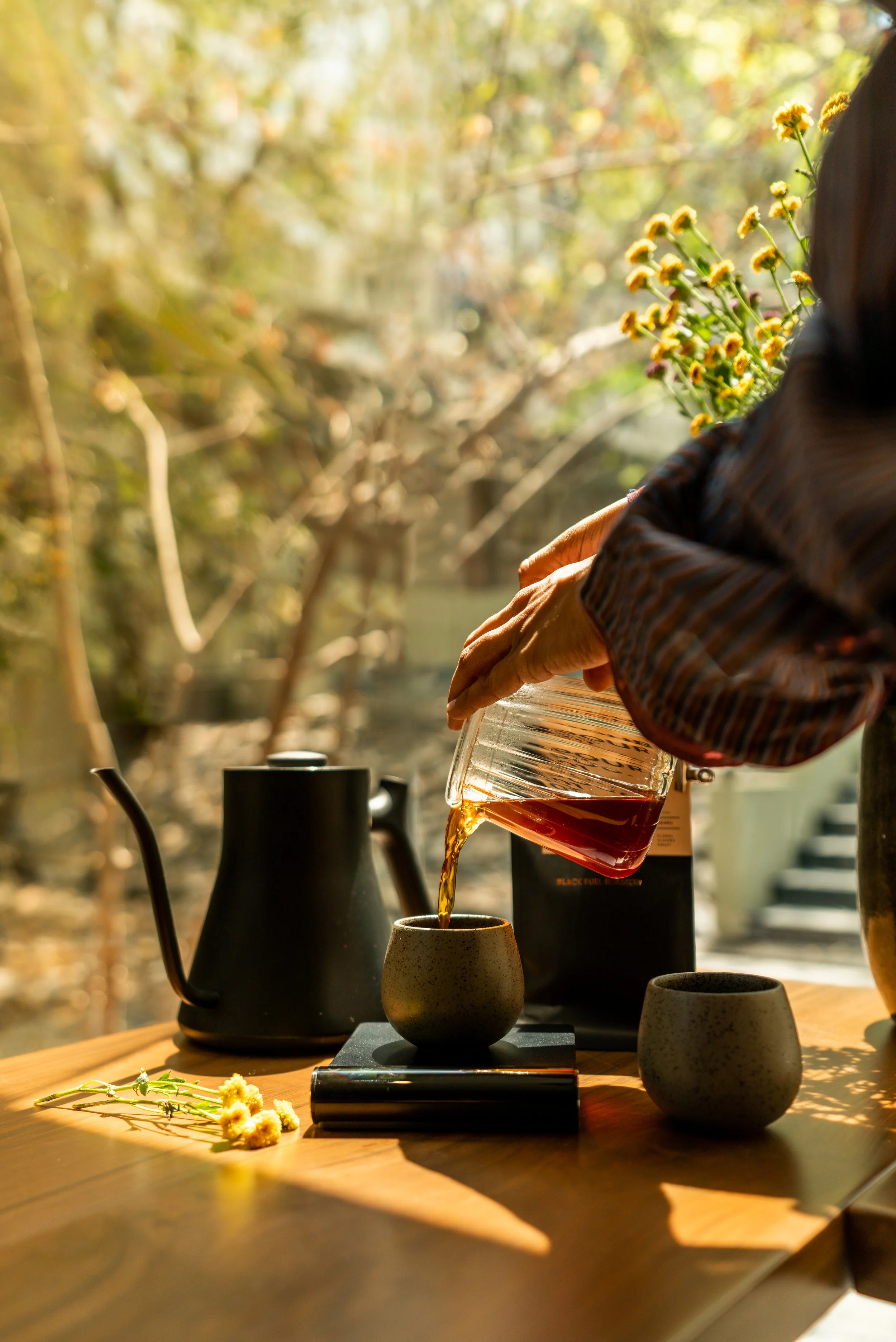 Someone pours hot tea from a glass pitcher into a ceramic cup on a wooden table, with a black teapot, another cup, and a package of tea in the background, set outdoors during daylight with trees and yellow flowers.