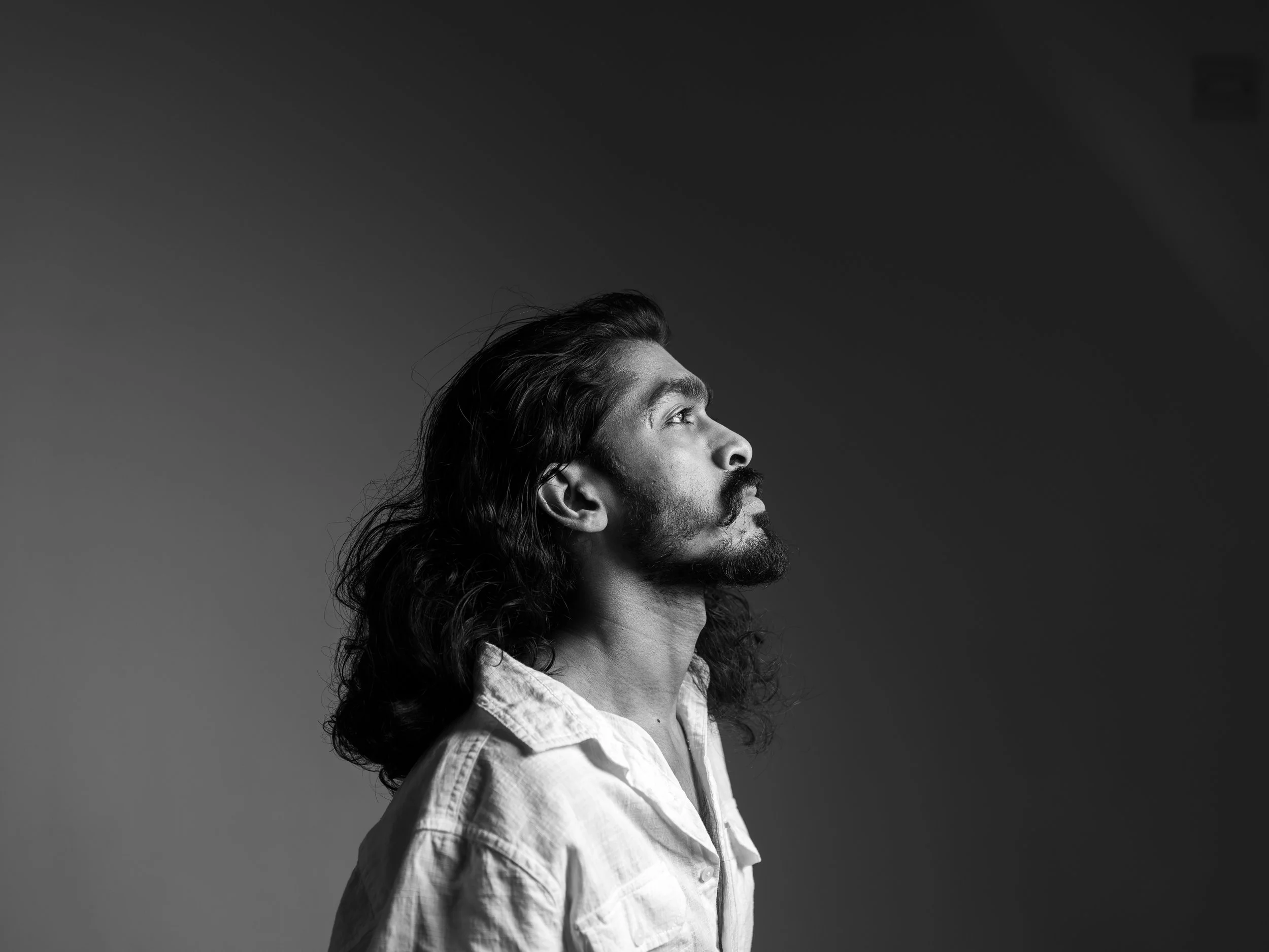 Black and white side profile of a man with long wavy hair and a beard, looking upward, wearing a light-colored shirt.