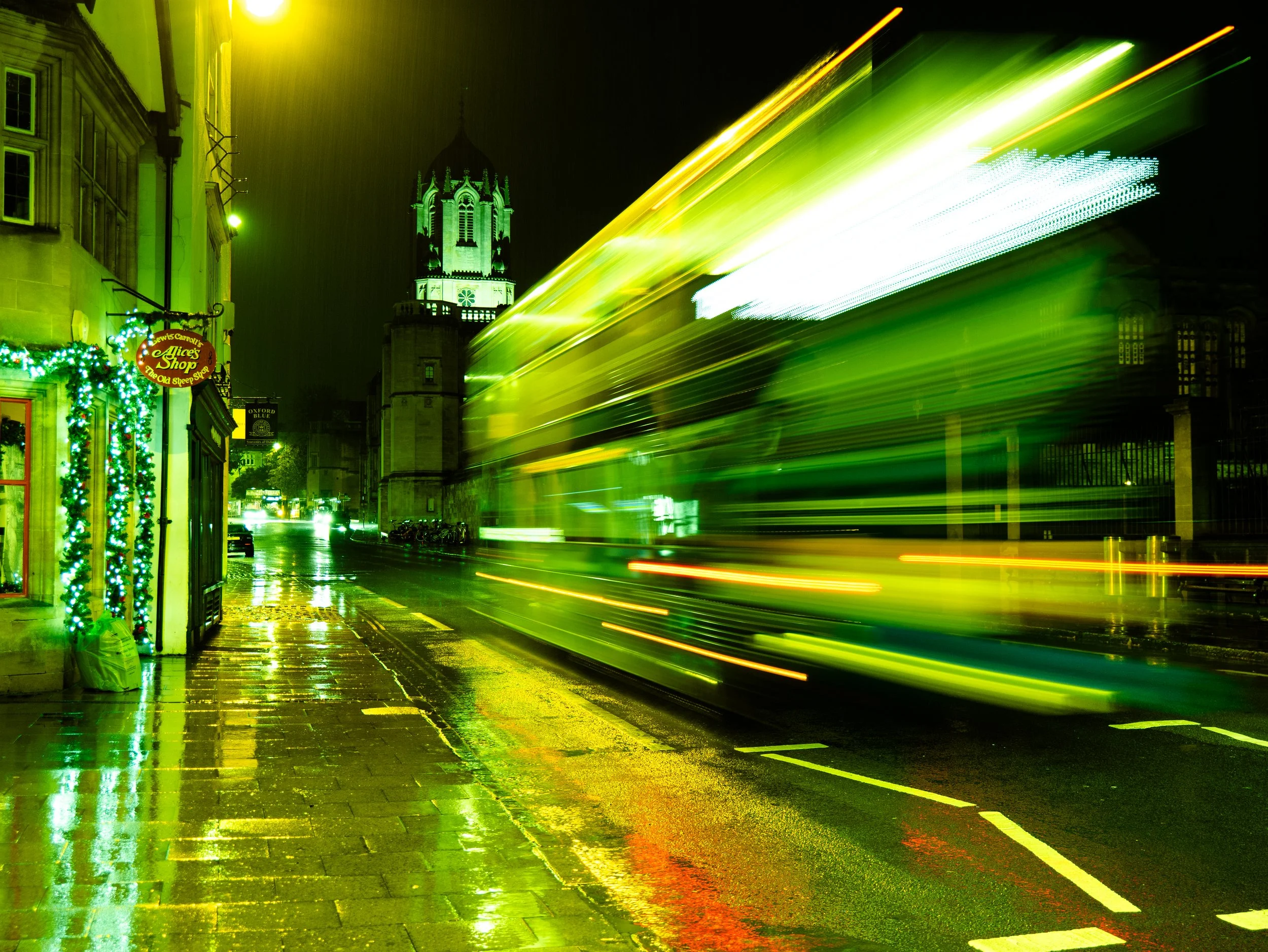 Night city street scene with colorful light trails from moving vehicles, wet pavement reflecting lights, a building with holiday decorations, and a church tower in the background.