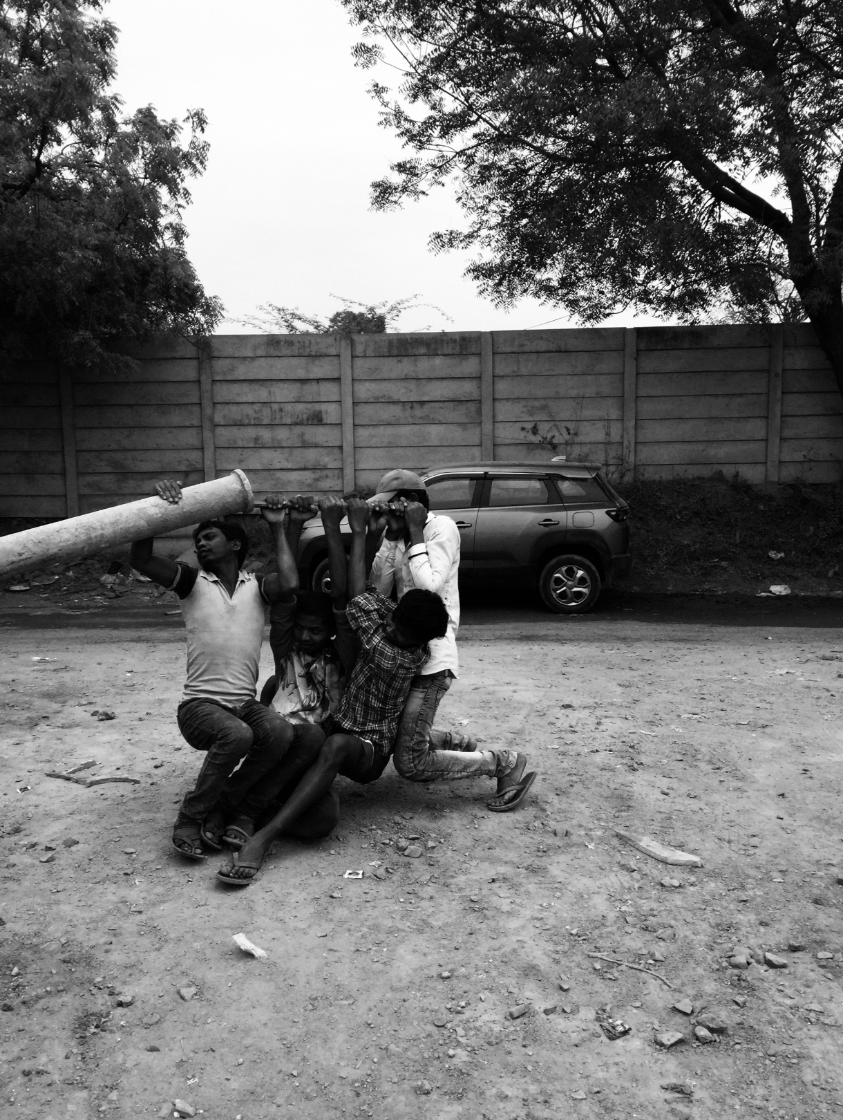 Four children playing with a large pipe on a dirt road, with a car and a wooden fence in the background.