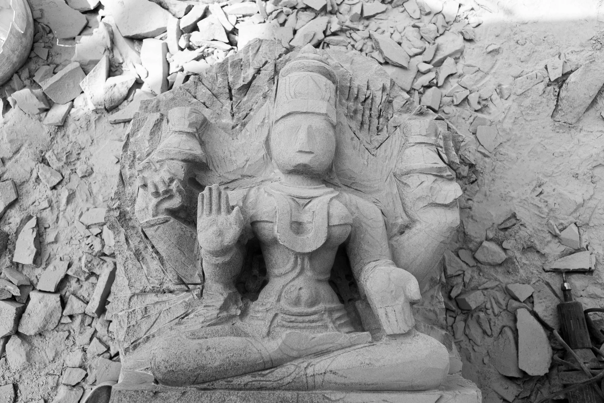 A carved stone sculpture of a Hindu deity, possibly Lord Vishnu, with a headdress, multiple arms, and jewelry, in progress with tools and rubble surrounding it.