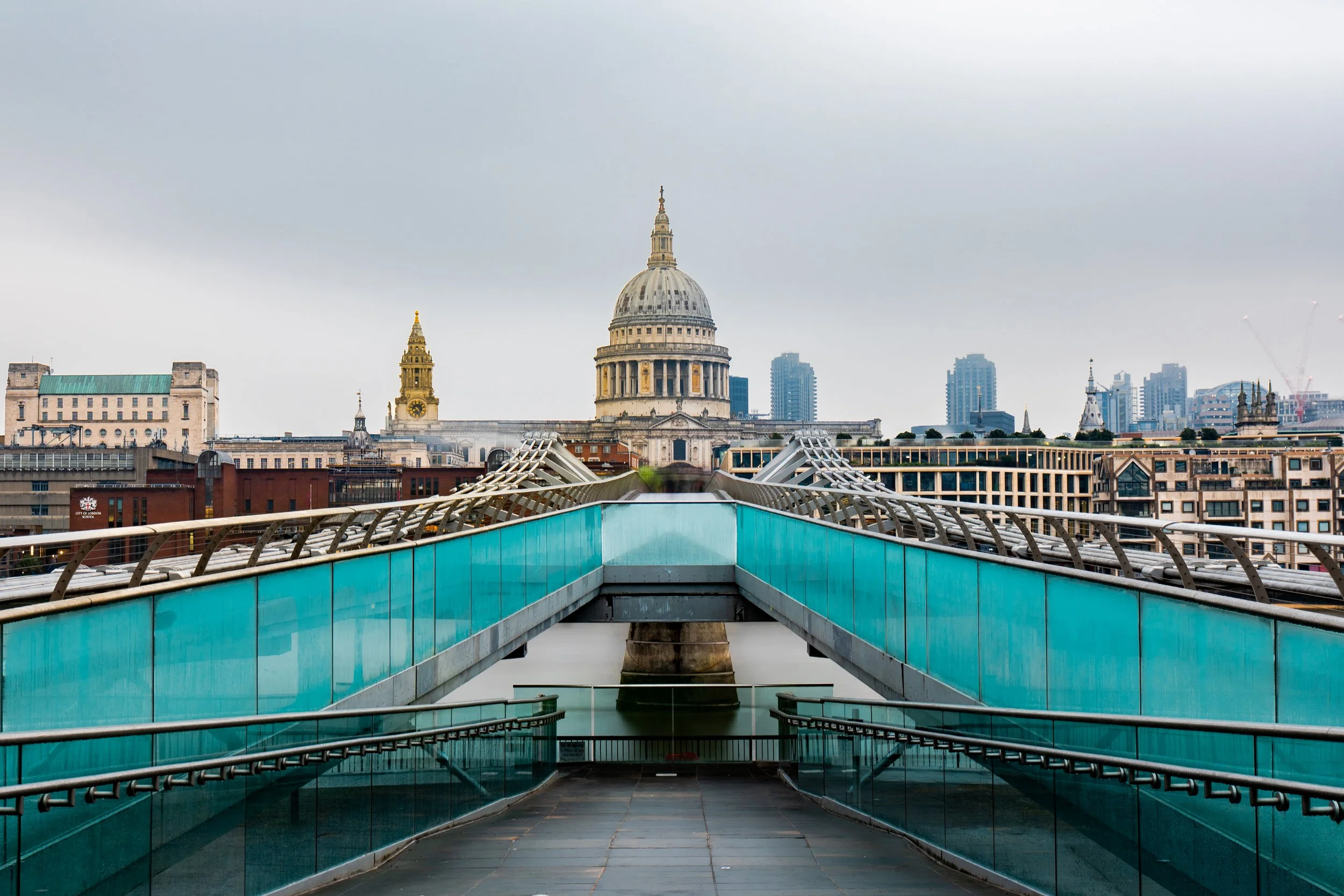 A modern glass and steel pedestrian bridge in London leading towards St. Paul's Cathedral, with city skyscrapers in the background on a cloudy day.