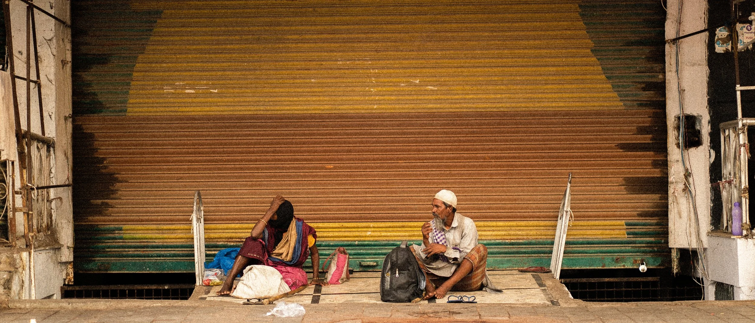 Two people sitting on the sidewalk in front of a closed, multi-colored, roll-up metal shutter.