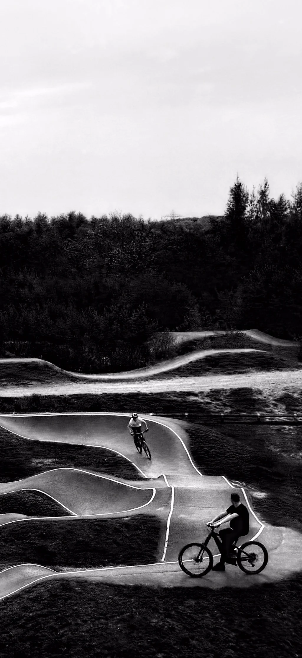 Two people riding bikes on a winding outdoor bike trail in a natural setting with trees and hills in the background.