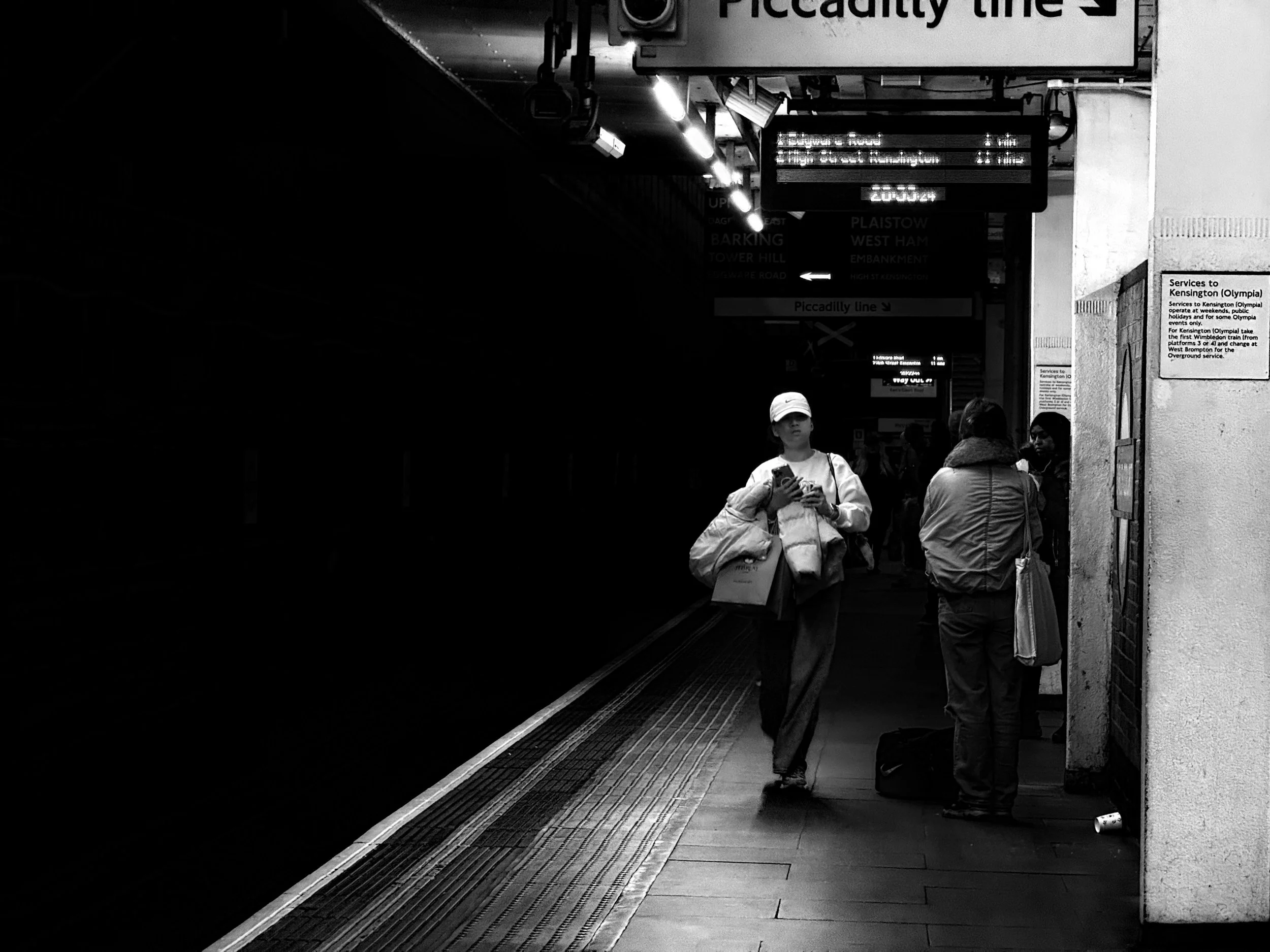 People waiting on a London Underground platform, with some looking at their phones. The platform is dimly lit, with signs indicating the Piccadilly line and train schedules.