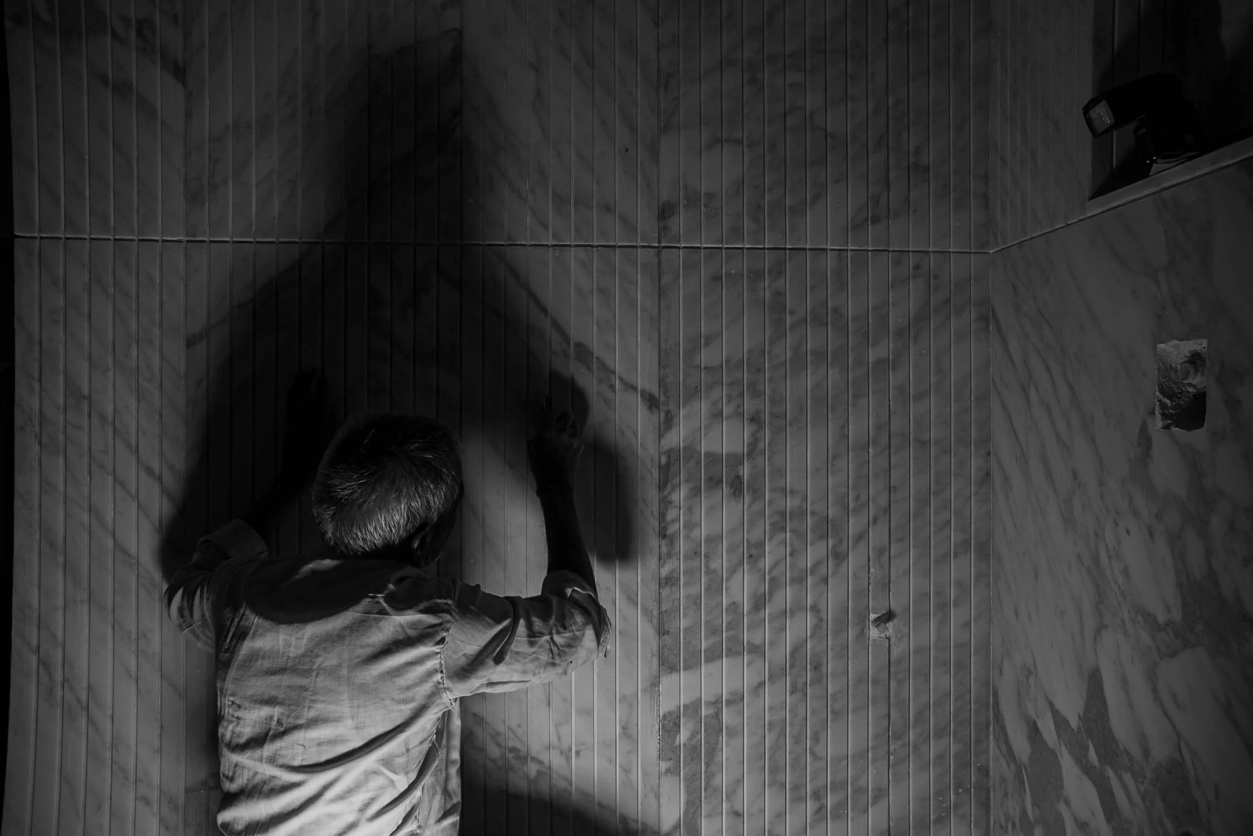 Top-down black and white photo of a man with short hair, seen from above, kneeling on a wooden floor in a corner of a room with tiled walls, working on the floor with his hands.
