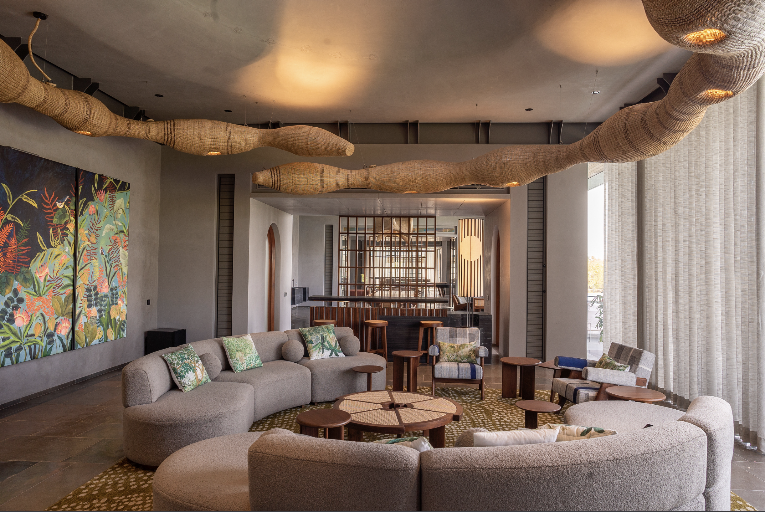 Interior of a modern hotel or lounge area with a curved beige sofa set, colorful cushions, small wooden tables, large sliding glass windows with curtains, artwork on the wall, and sculptural fabric installations hanging from the ceiling.