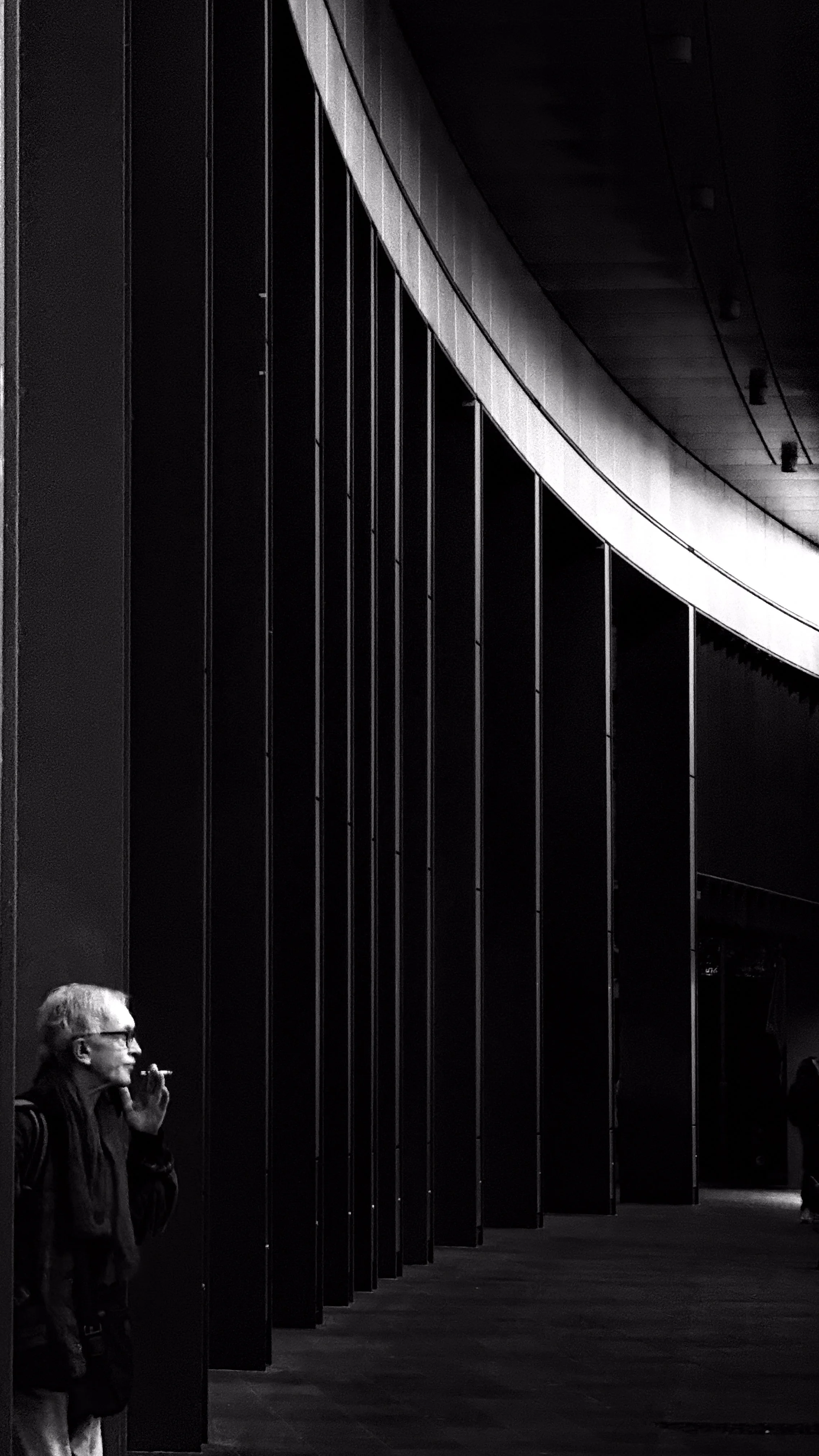 A woman with glasses smoking a cigarette stands next to a large modern building with dark vertical metal panels.
