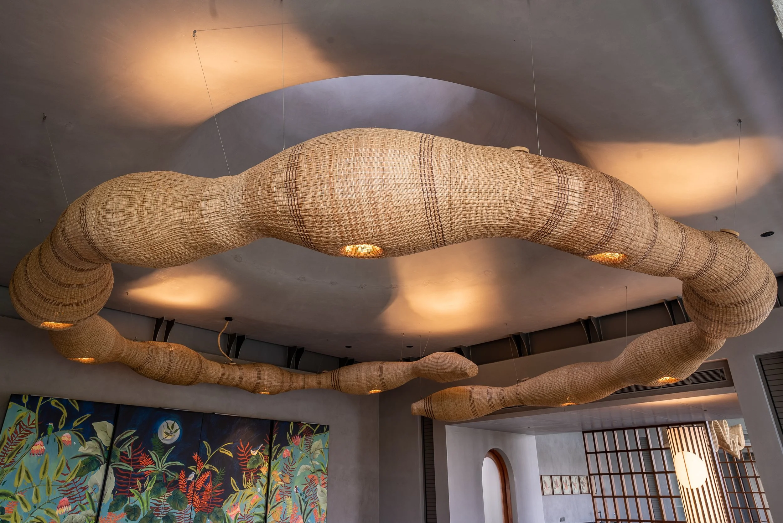 Unique ceiling light fixture made of woven natural fibers in organic, flowing shapes, installed in a modern interior space with artwork on the wall.