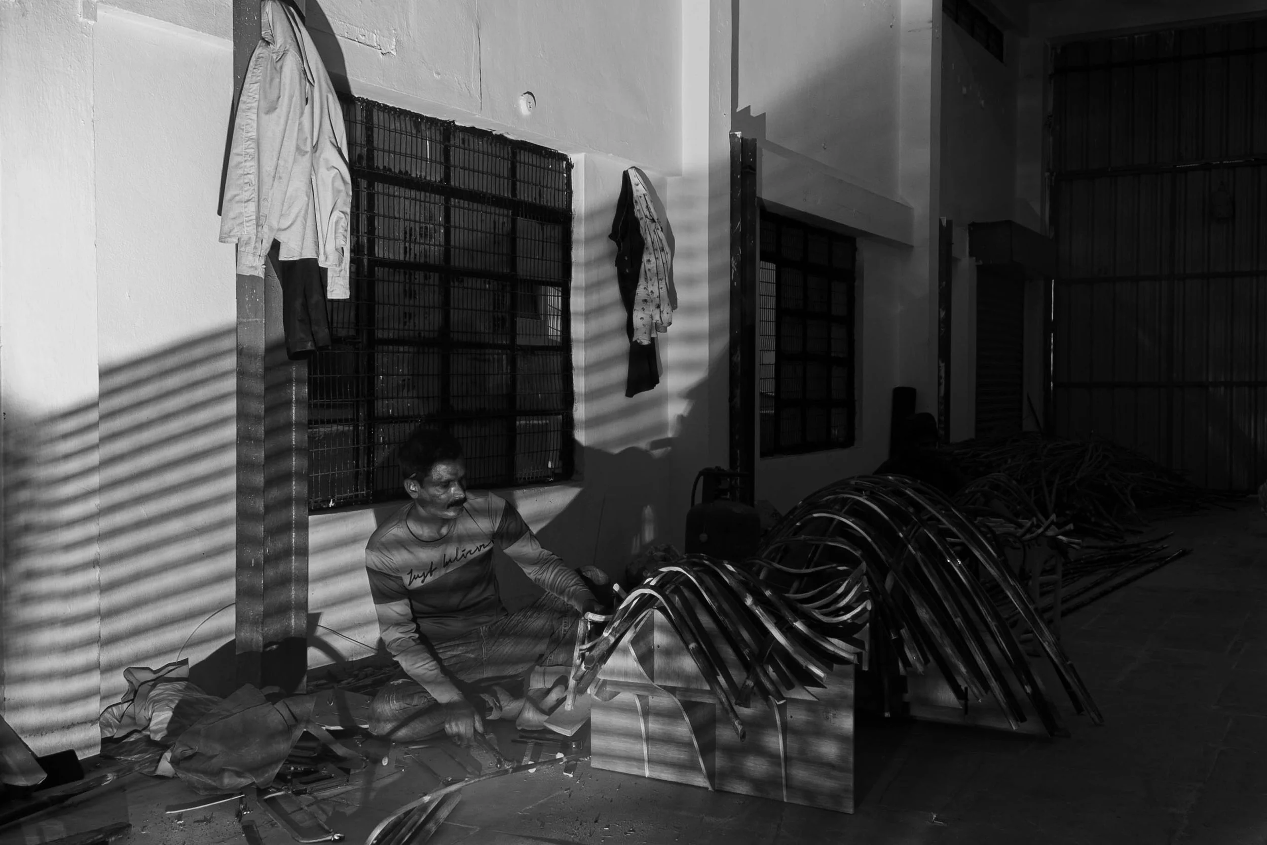 A man sitting on the floor in a room with construction materials, metal frames, and hanging clothes, with shadows cast by window blinds.