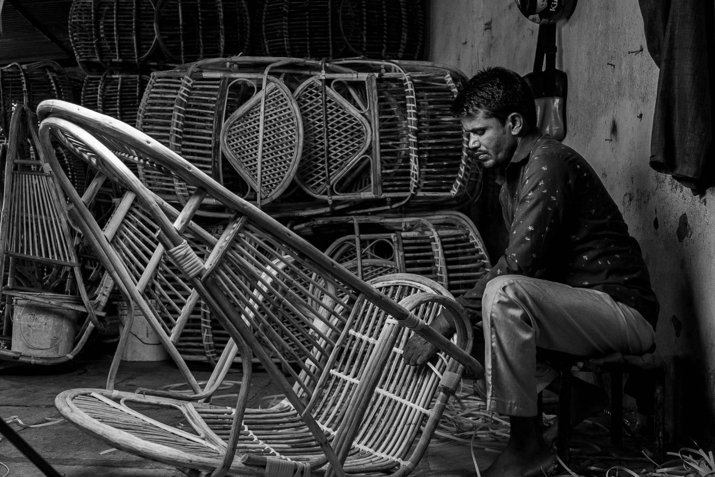 A man sitting on a stool in a workshop, surrounded by stacks of rattan furniture and chairs in various stages of production.