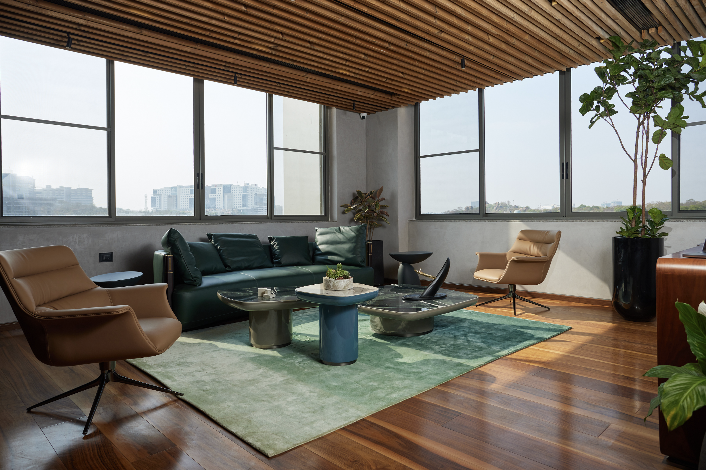 Modern living room with large windows, brown and green leather chairs, a green sofa, a marble-top coffee table, green area rug, tall potted plants, and wooden ceiling beams.