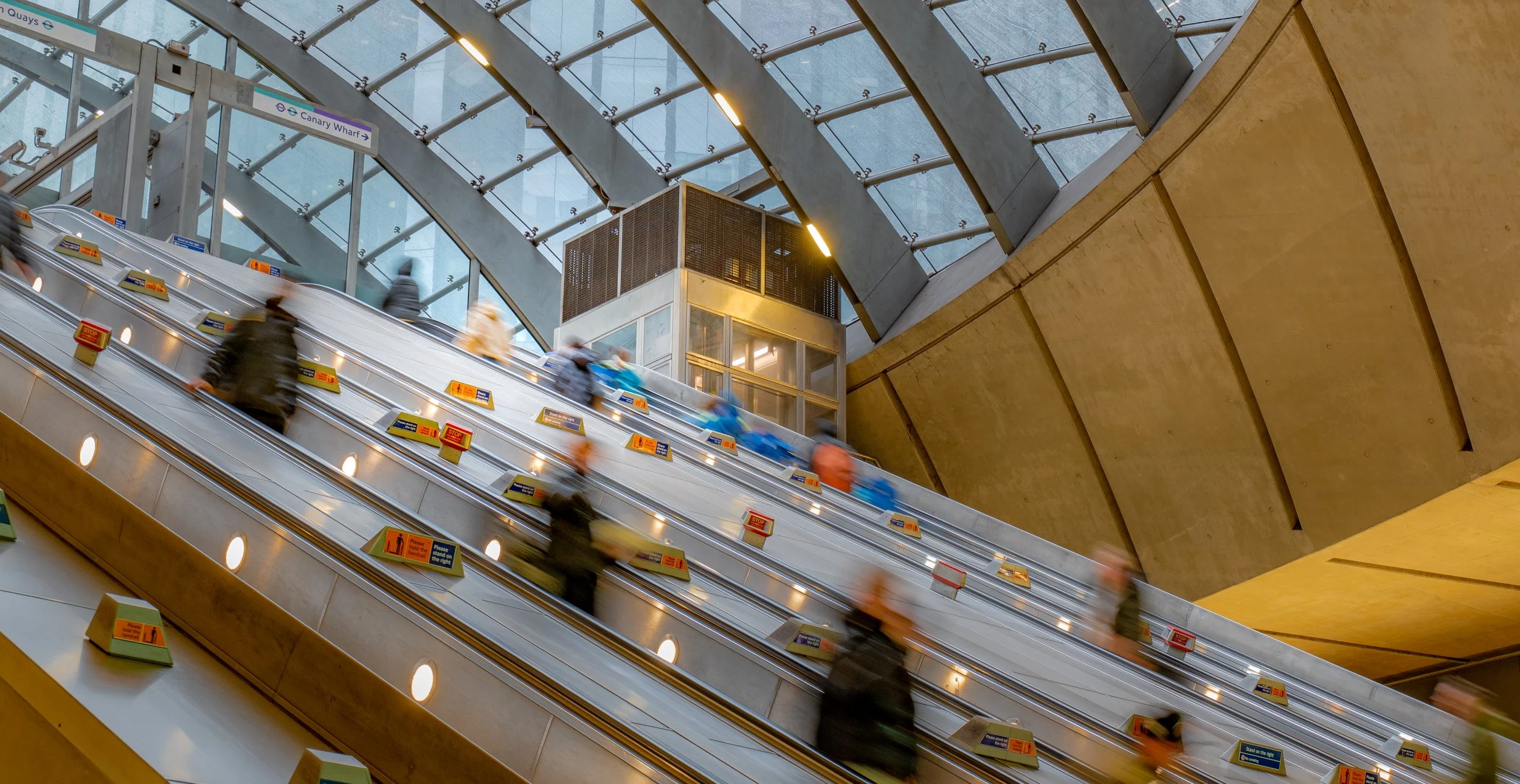 London Underground station escalators with passengers moving up and down, sign indicating Canary Wharf, modern glass and metal architecture, and yellow wall panels.