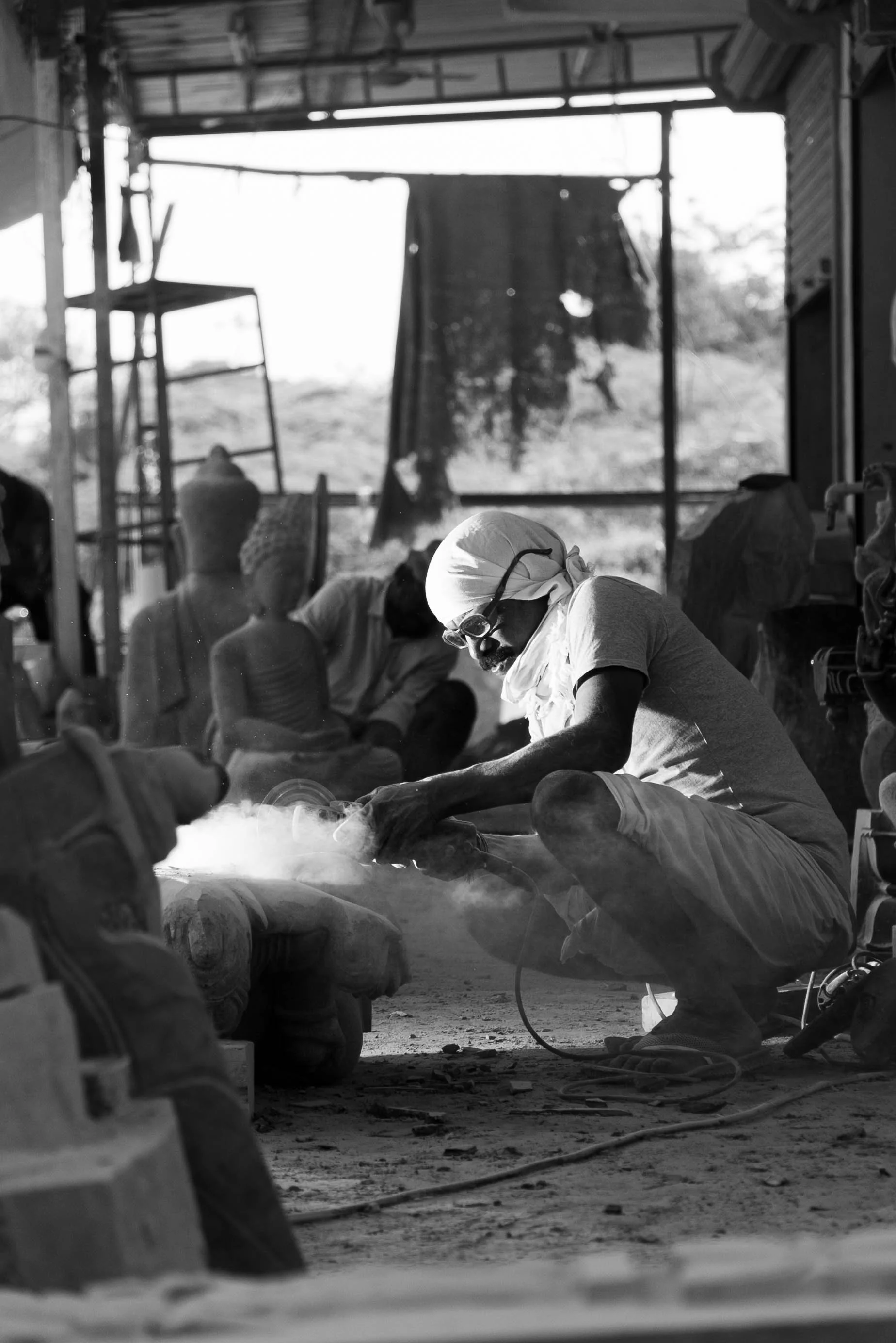 A man wearing a turban and glasses is welding or grinding, creating smoke in a workshop with other people working in the background.