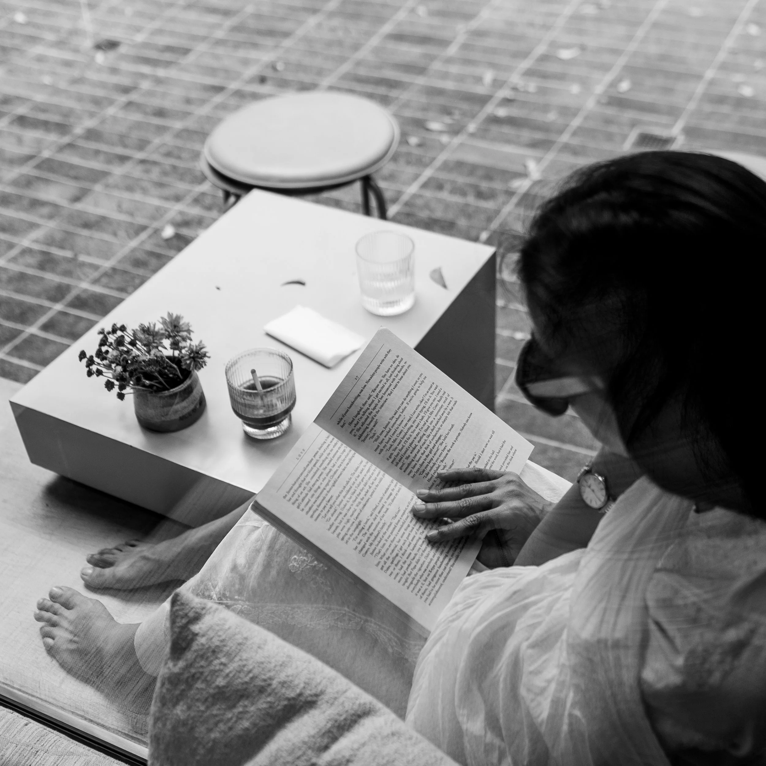 A person sitting indoors reading a book, with a small table beside them holding two glasses of water, a small potted plant, and a candle, viewed from above.