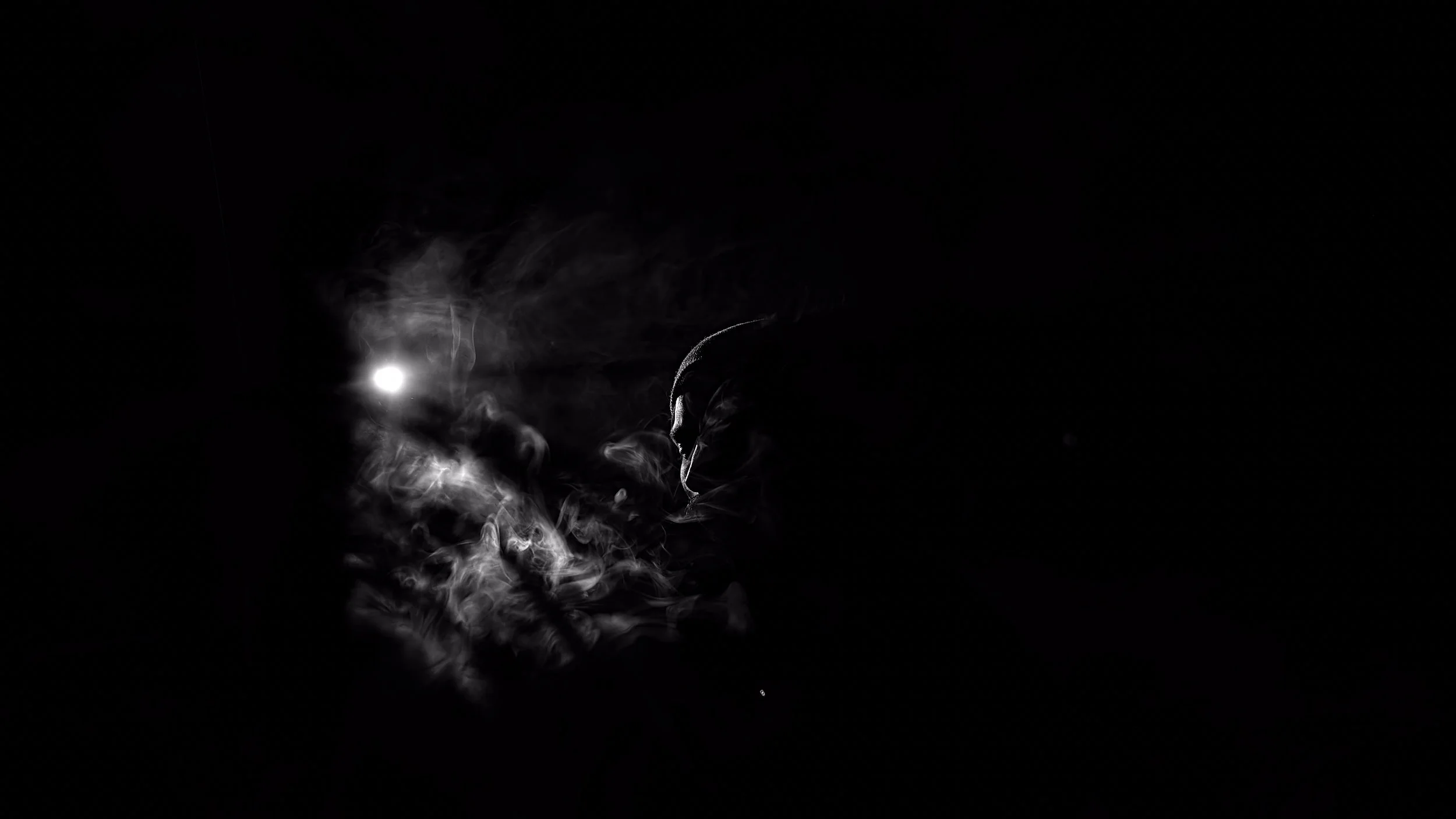 A dark image with a bright light source in the upper left, creating smoke-like wisps and a silhouette of a person wearing a hat or cap in the lower right.