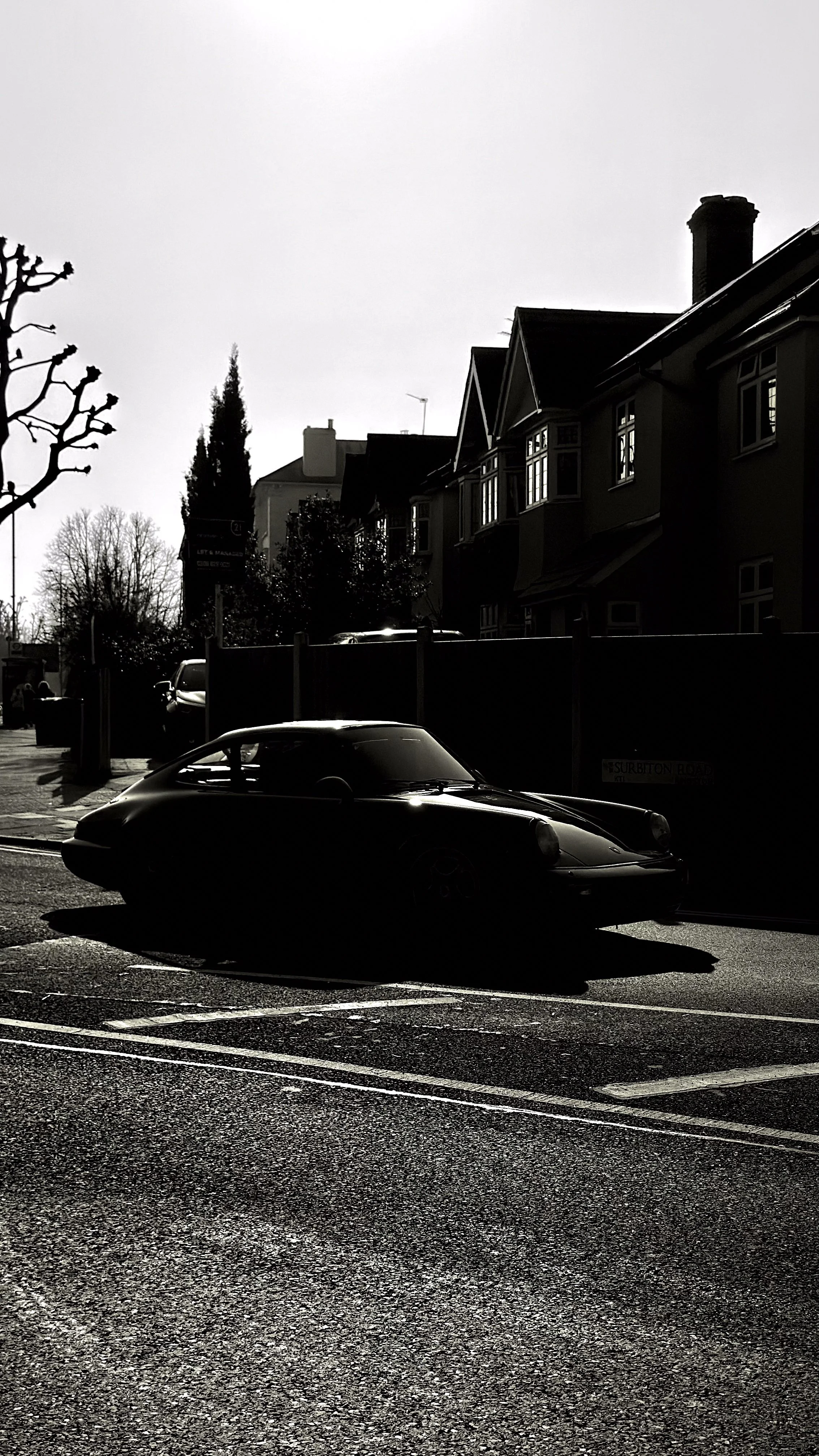 A black vintage sports car parked on the street with houses and trees in the background, backlit by the bright sky.