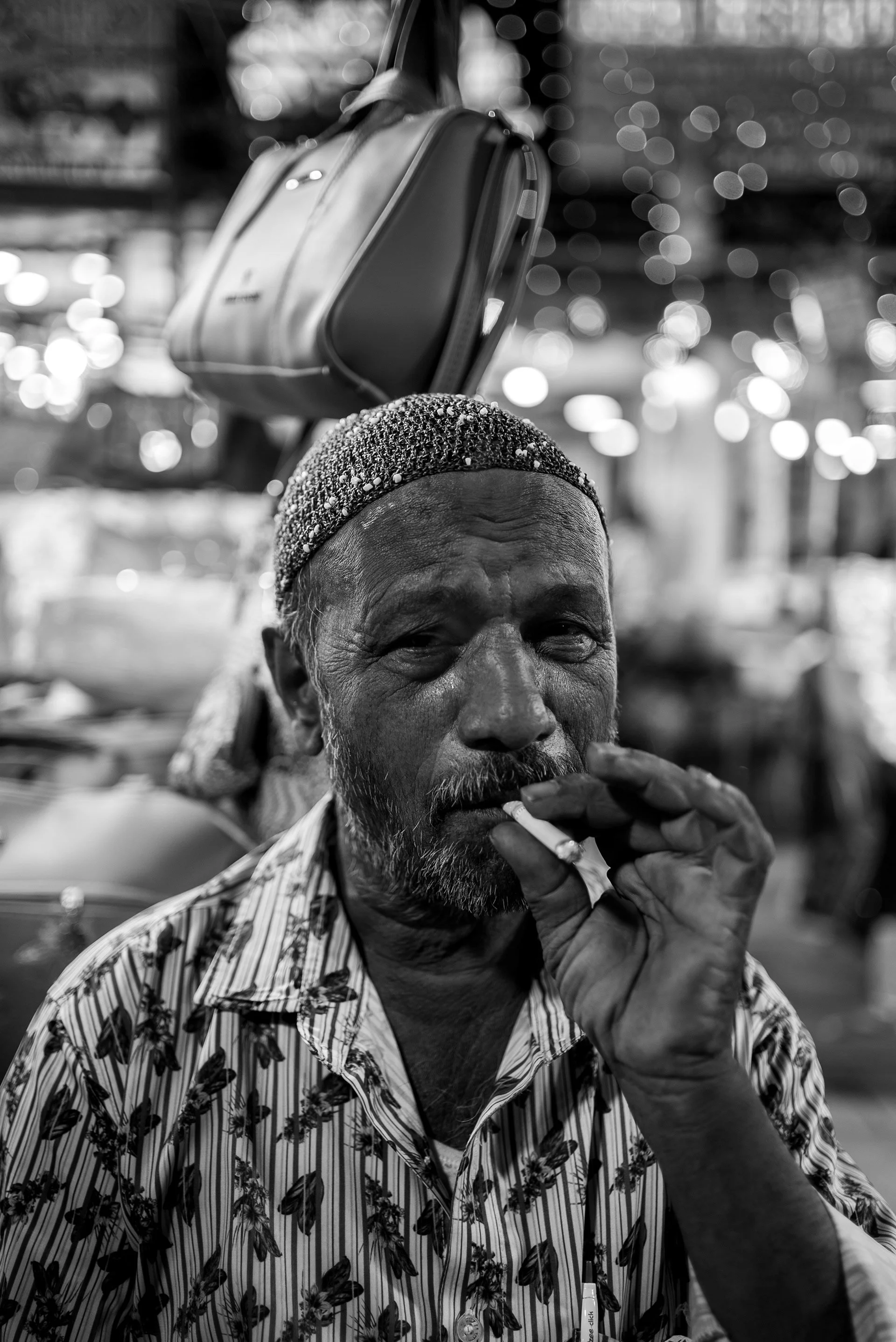 A man with a beard and patterned shirt smokes a cigarette in a busy, illuminated market. A bag hangs above him, and colorful lights create a bokeh background.