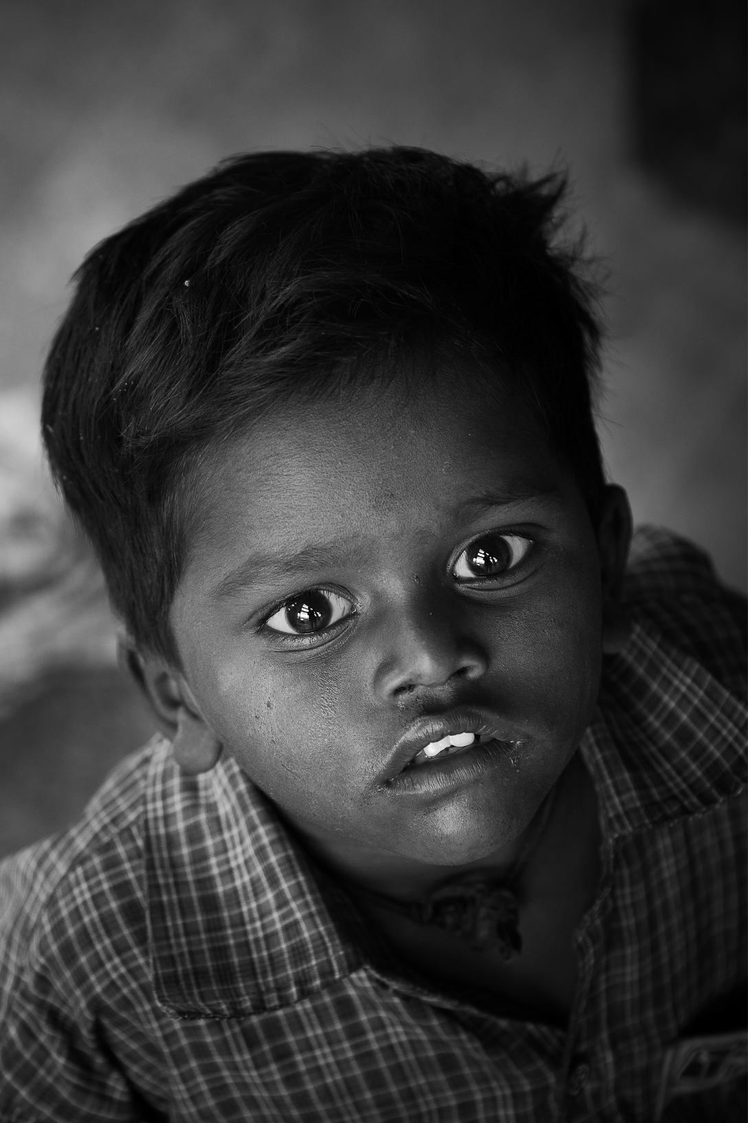 Black and white close-up portrait of a young boy looking directly at the camera with wide eyes and slightly parted lips, wearing a checkered shirt.