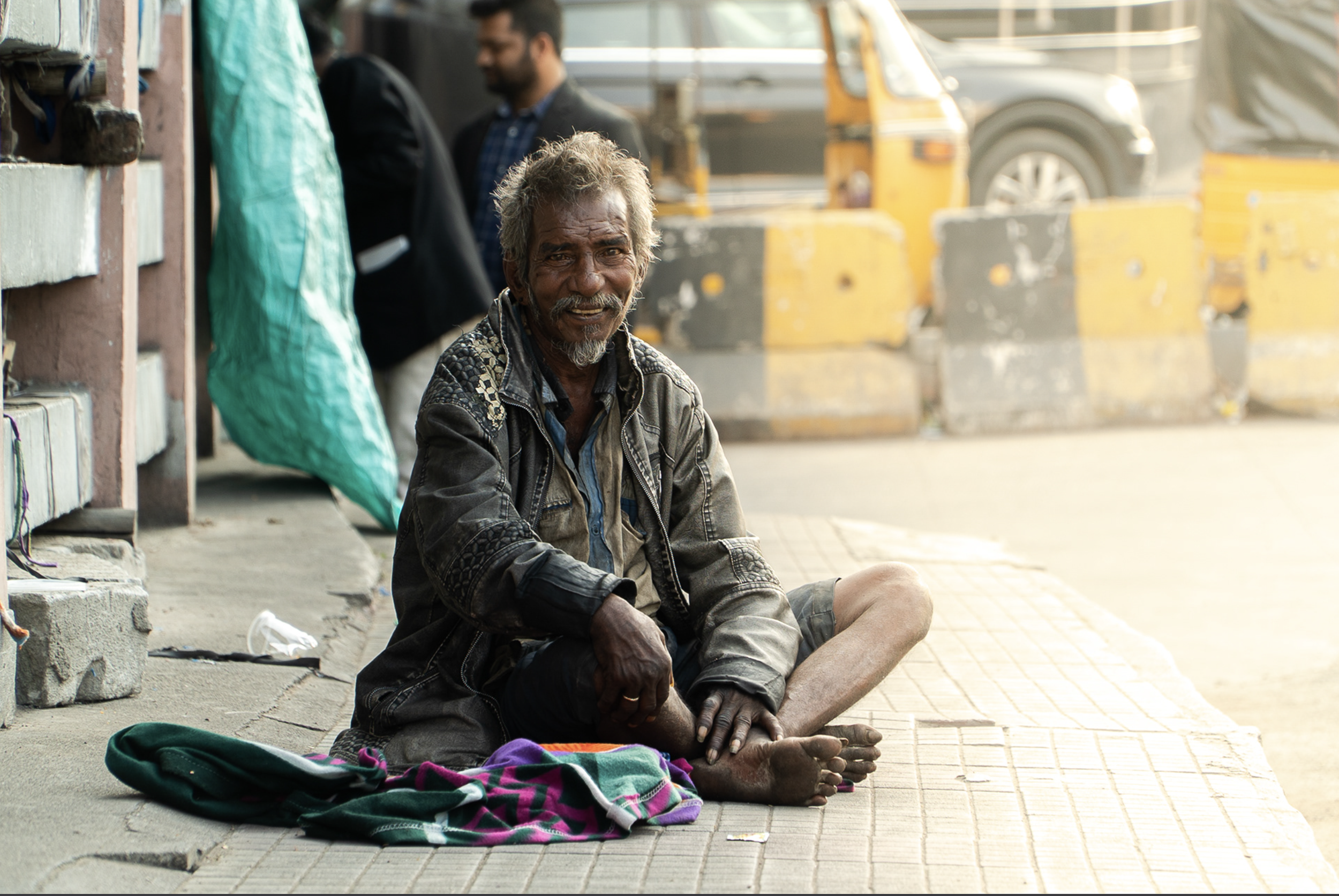A homeless man sitting on the sidewalk with a colorful blanket, wearing a dark jacket and shorts, looking at the camera with a slight smile.