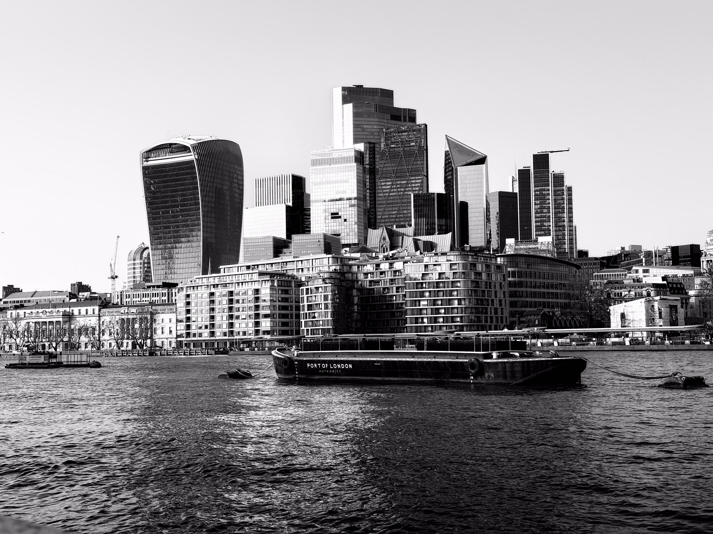 Black and white photo of a city skyline with tall modern skyscrapers and buildings near a river. A boat labeled 'Port of London' is visible on the water in the foreground.