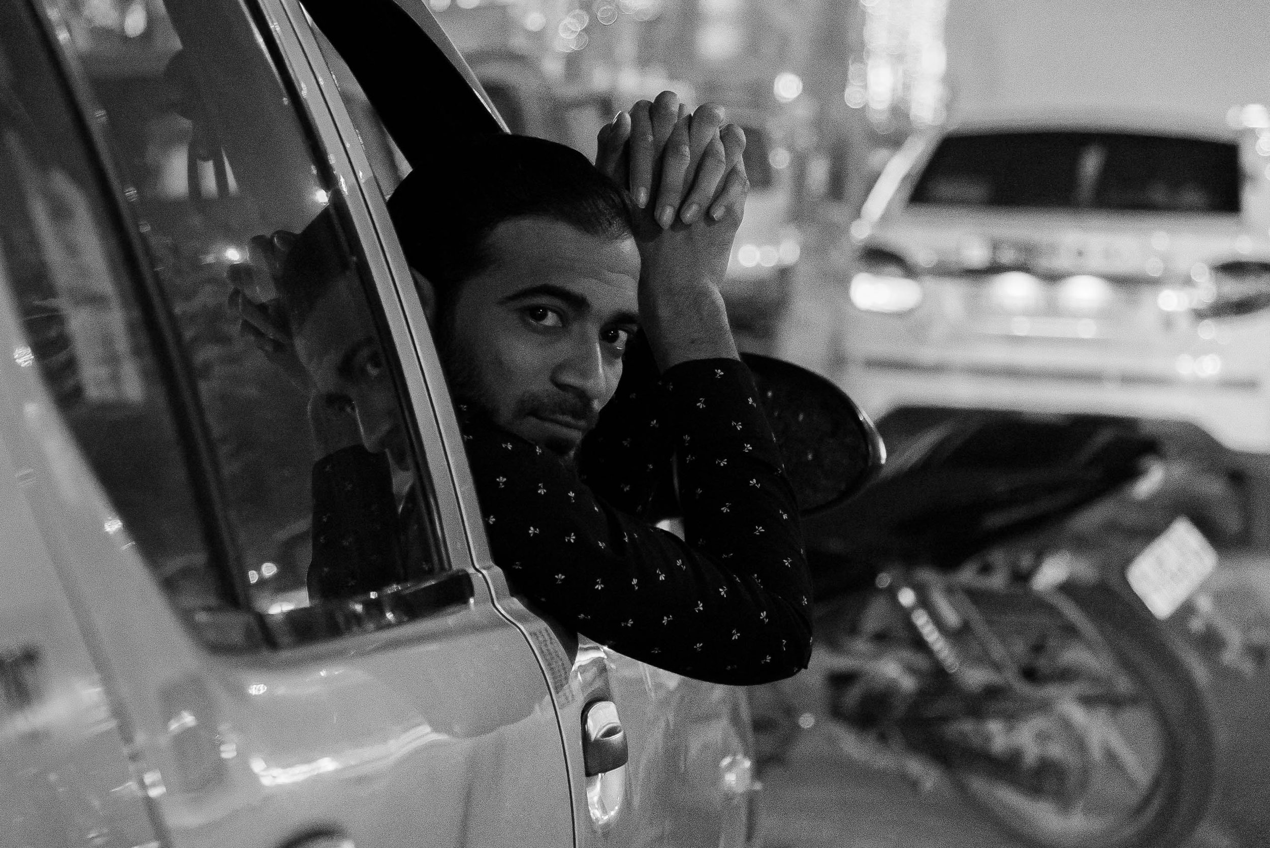A man with dark hair and a beard, wearing a dark shirt with small patterns, leaning out of a vehicle's window, looking at the camera, with a blurry background of parked vehicles and a motorcycle.