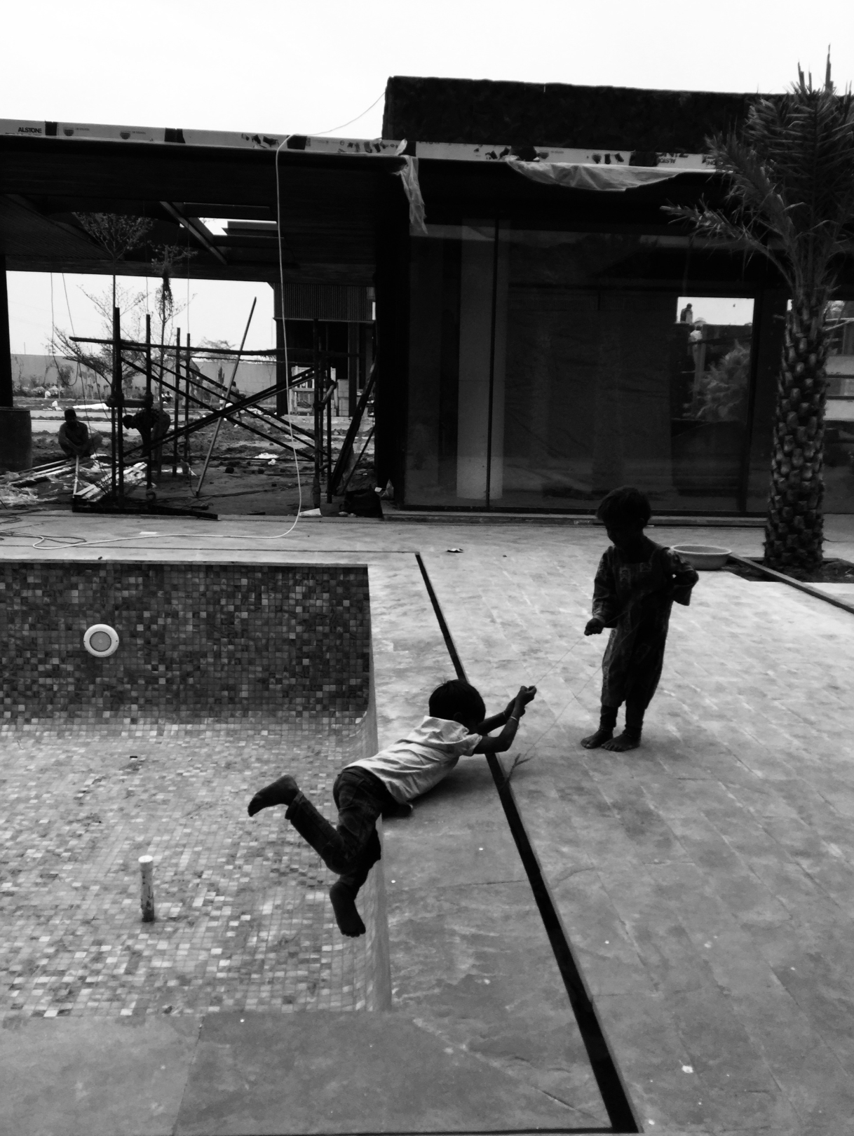Two children playing near a swimming pool on a construction site, with people working in the background and a building under construction.