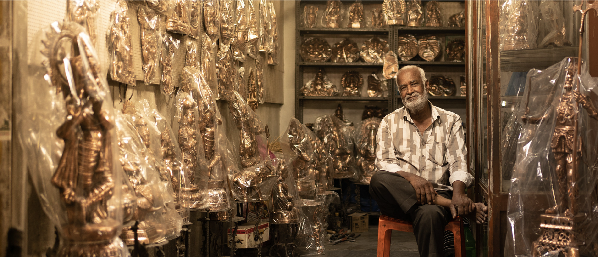 An elderly man with a beard sitting on a stool inside a shop filled with metallic Indian statues wrapped in plastic. Shelves and walls are lined with various statues, mostly of gods and deities, in a warm, dimly lit environment.