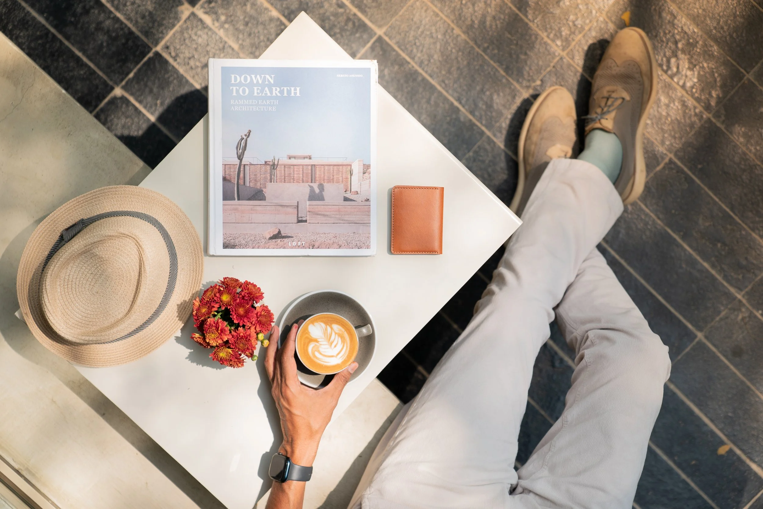 Overhead view of a person sitting at a white table with a cappuccino, a sun hat, a small potted flower, a magazine titled "Down to Earth," a leather wallet, and a cup of coffee art. The person is wearing beige pants, gray shoes, and a smartwatch.