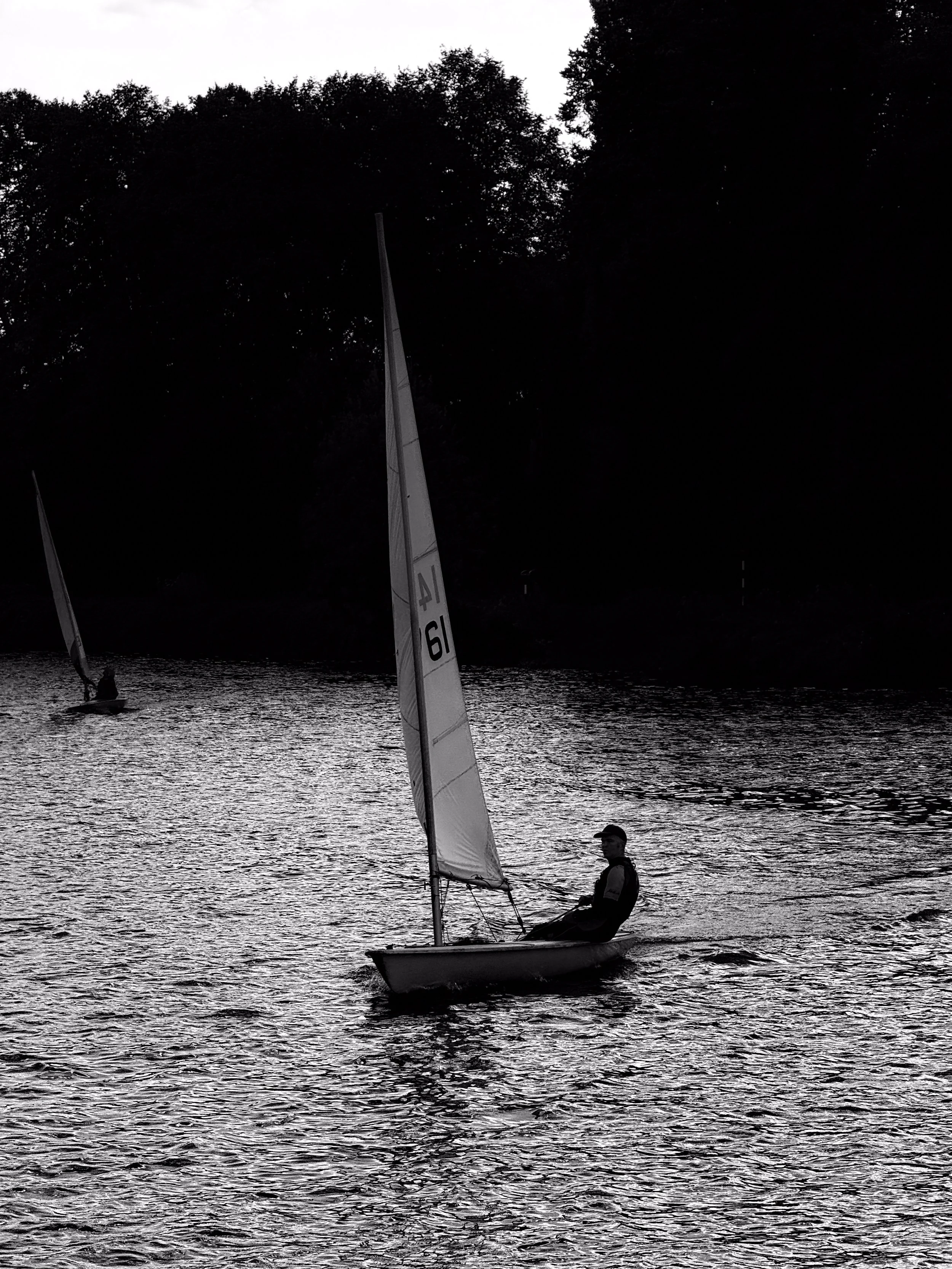 Two people sailing small boats on a body of water during the daytime, with a tree-lined shoreline in the background.