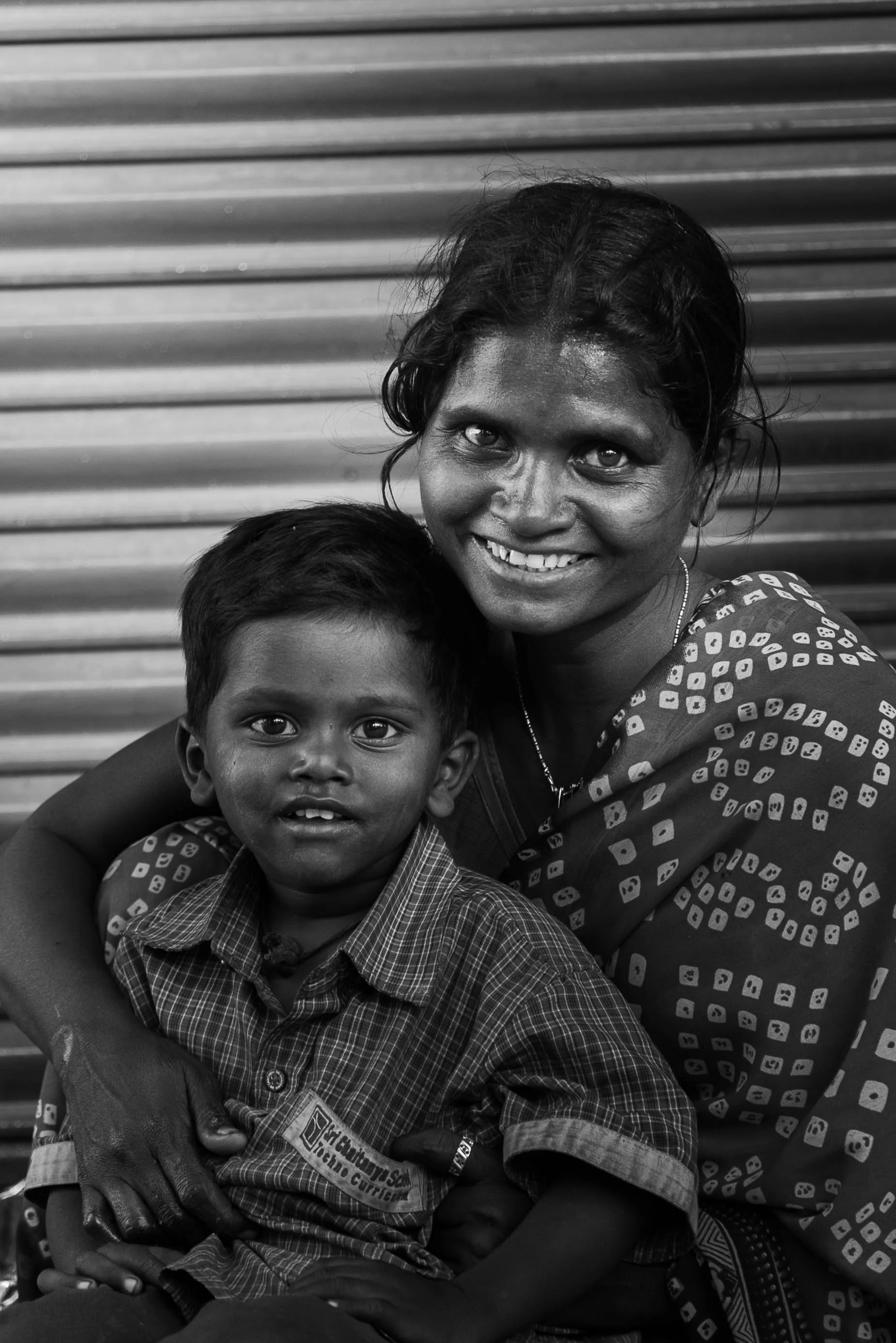 A smiling woman and a young boy sitting together, with the woman embracing him, against a background of horizontal metal shutters.