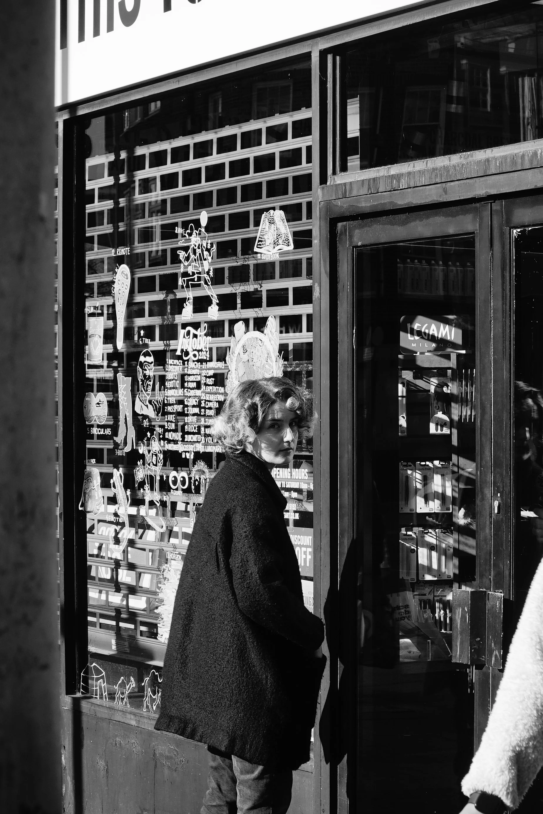 A woman with curly hair wearing a dark coat, standing outside a storefront with colorful stickers and signs, looking at the camera in a city setting.