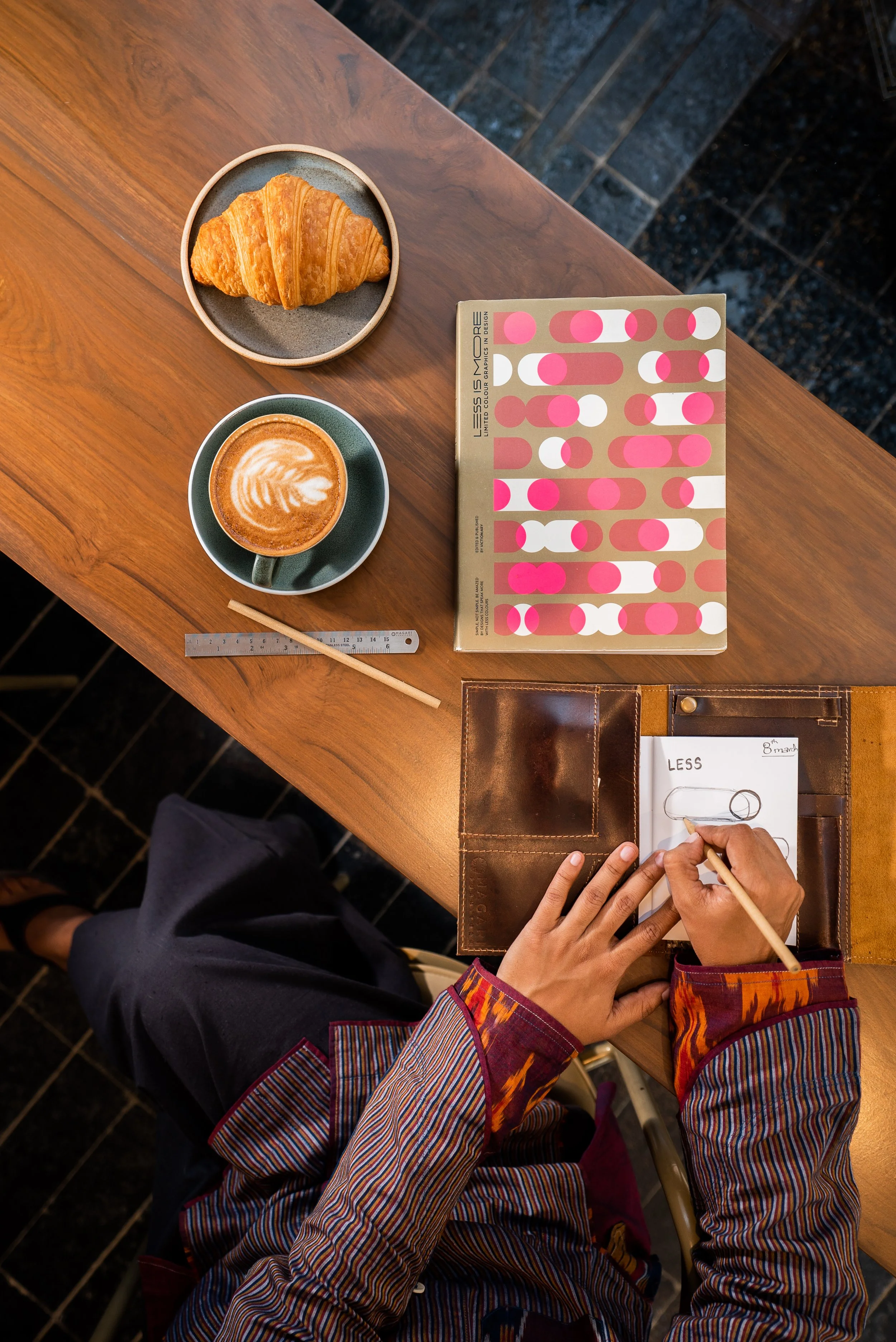 Top-down view of a wooden table with a croissant on a small plate, a cup of latte with latte art, a ruler, a colorful hotspot book, and a person sketching design ideas in a leather notebook.