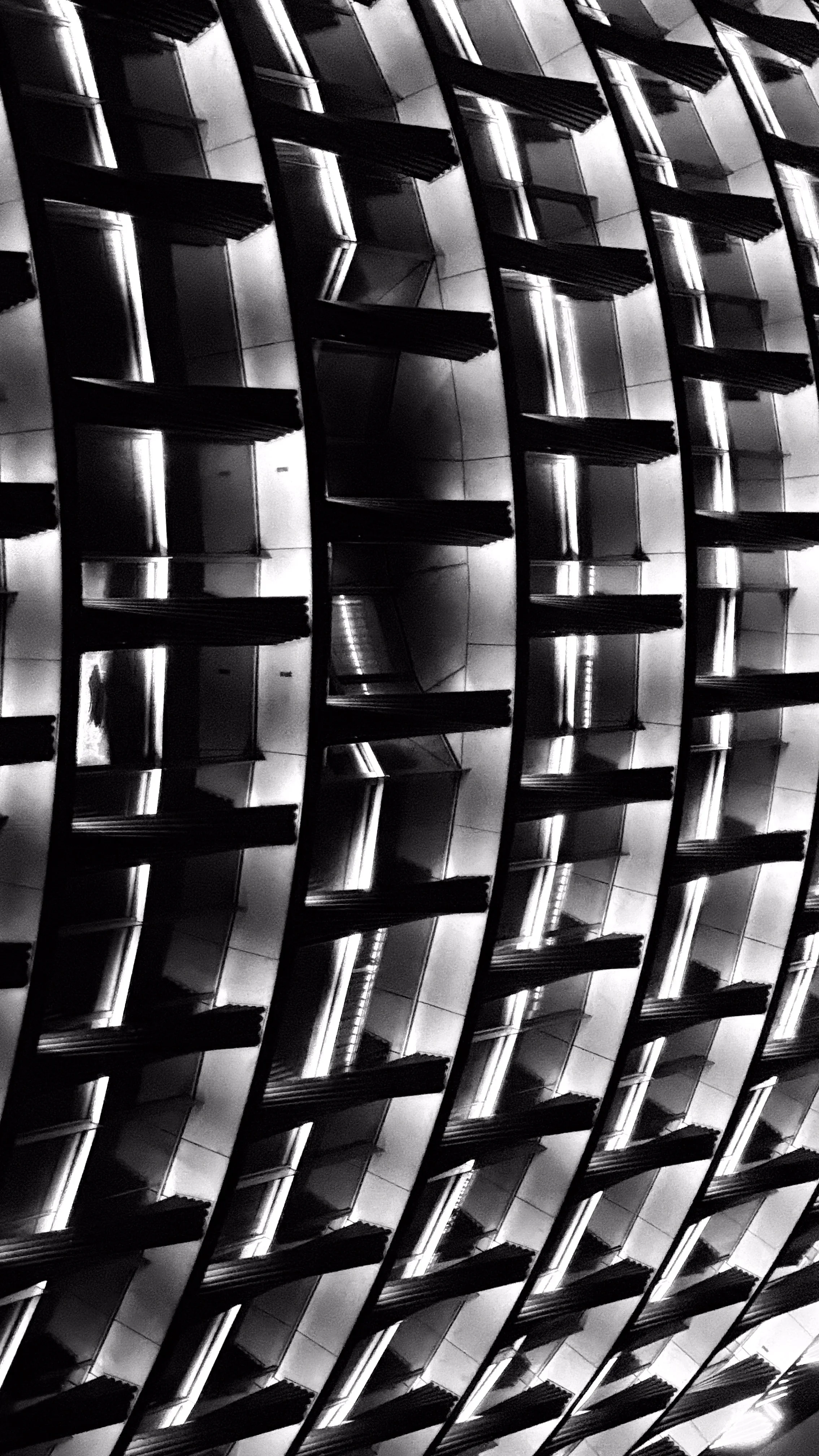 A high-angle view of a modern building facade with reflective windows and awnings, arranged in a repeating diagonal pattern, in black and white.