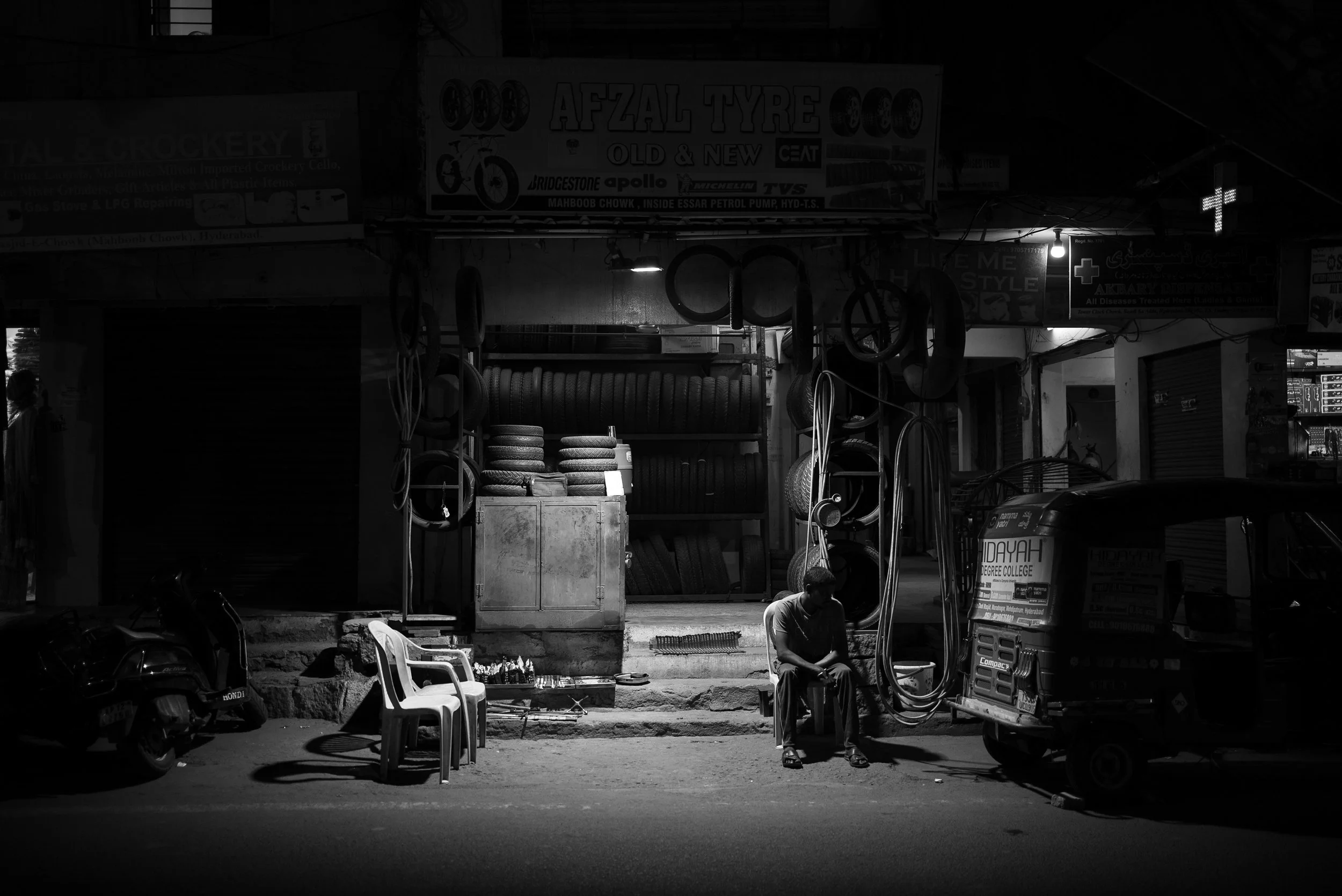 A man sitting on a chair in front of roadside tire shop at night with tires stacked behind him and a parked auto rickshaw nearby.