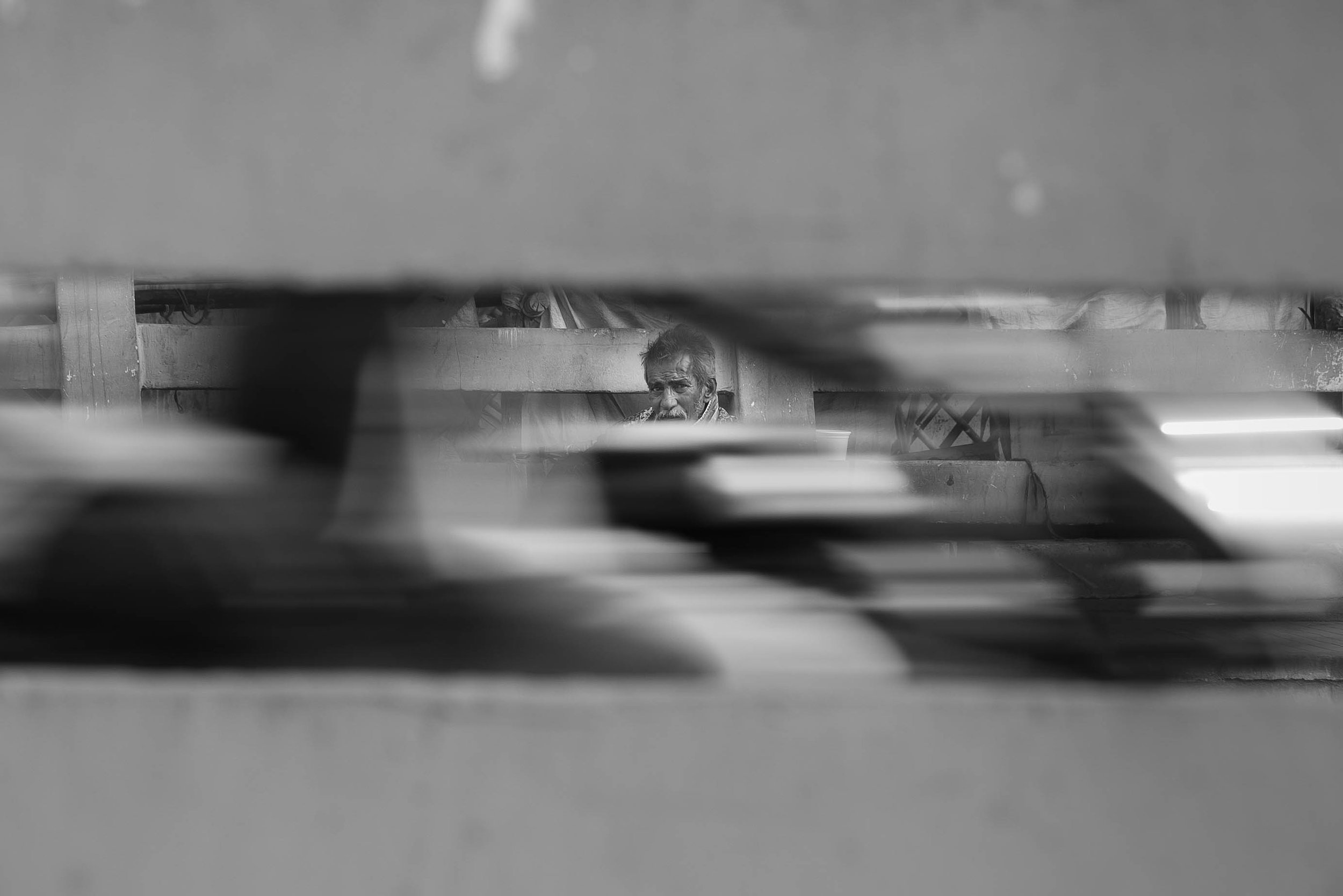 A man with a beard and disheveled hair sitting behind a structure, possibly a bench or platform, seen through a narrow gap. The photo is in black and white.