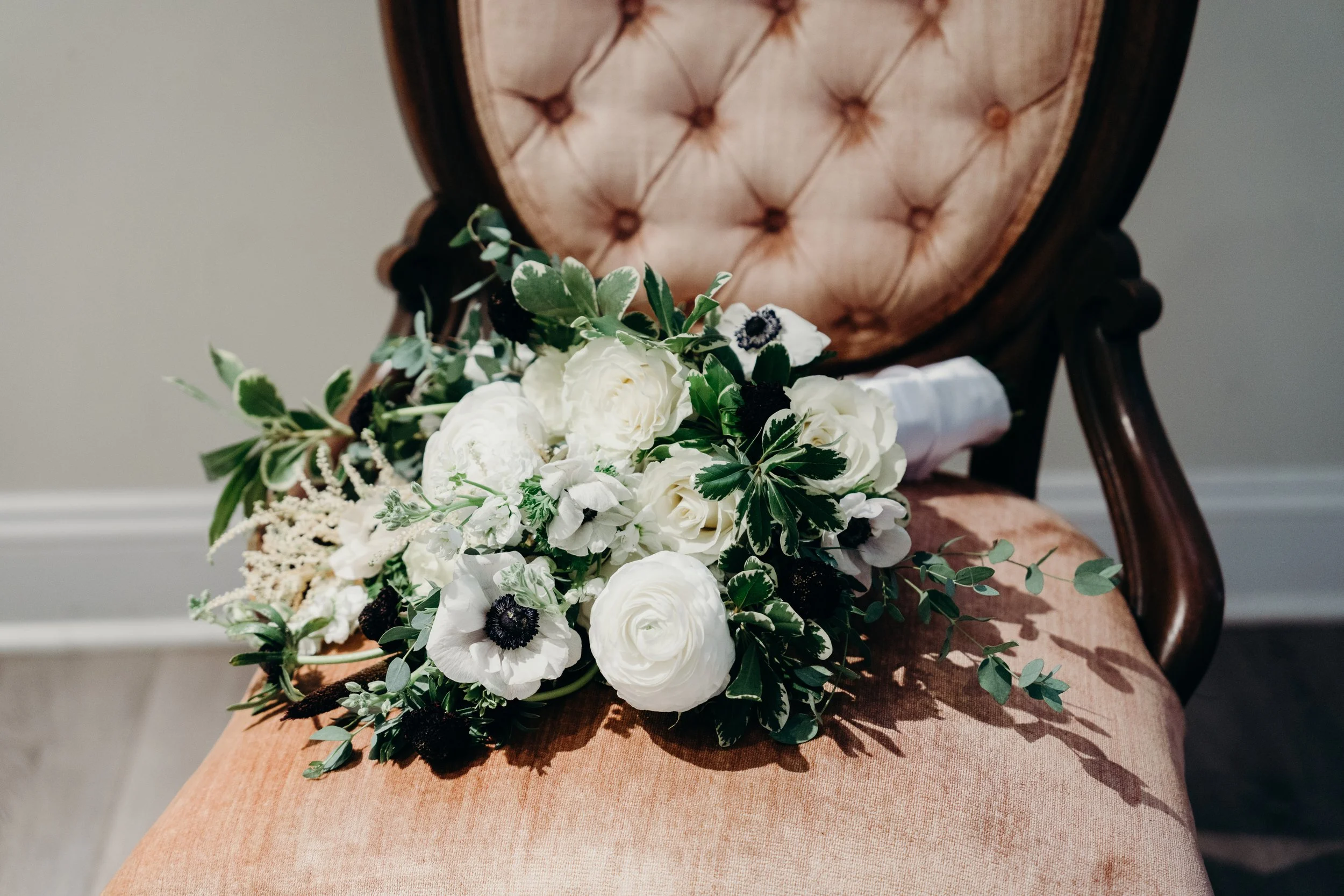 A bouquet of white flowers with green foliage resting on a vintage upholstered chair with tufted fabric and dark wooden frame.