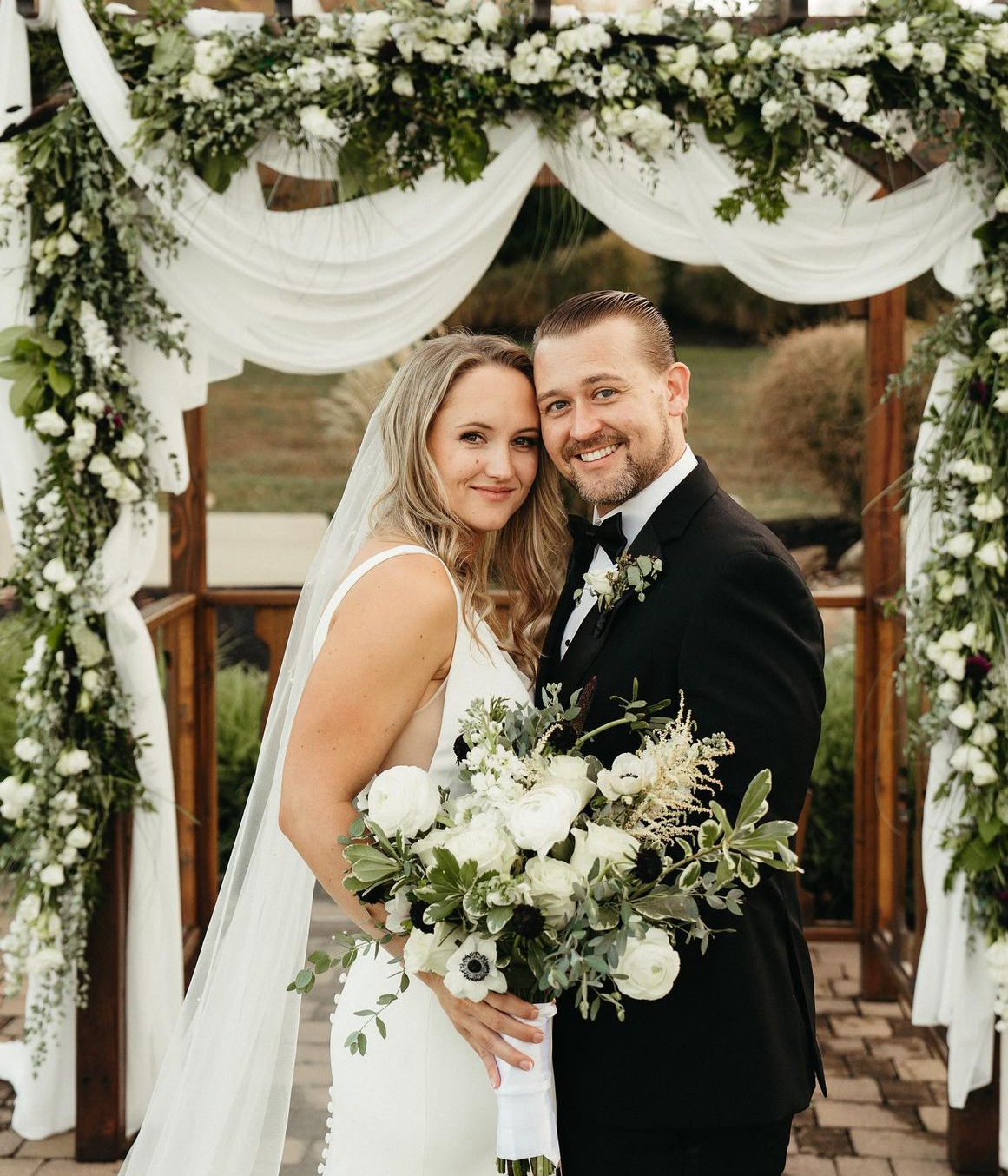A bride and groom standing together under a floral wedding arch, smiling at the camera, with the bride holding a large bouquet of white and green flowers.