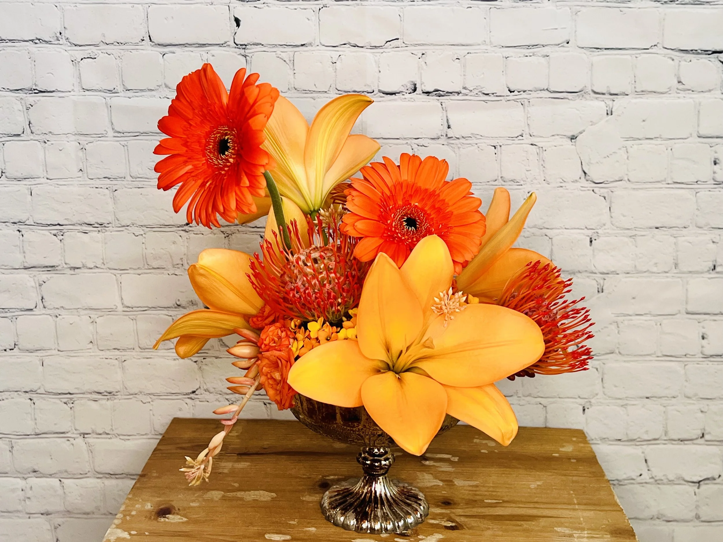 Arrangement of orange gerbera daisies, yellow lilies, and other flowers in a decorative silver vase on a wooden table against a white brick wall background.