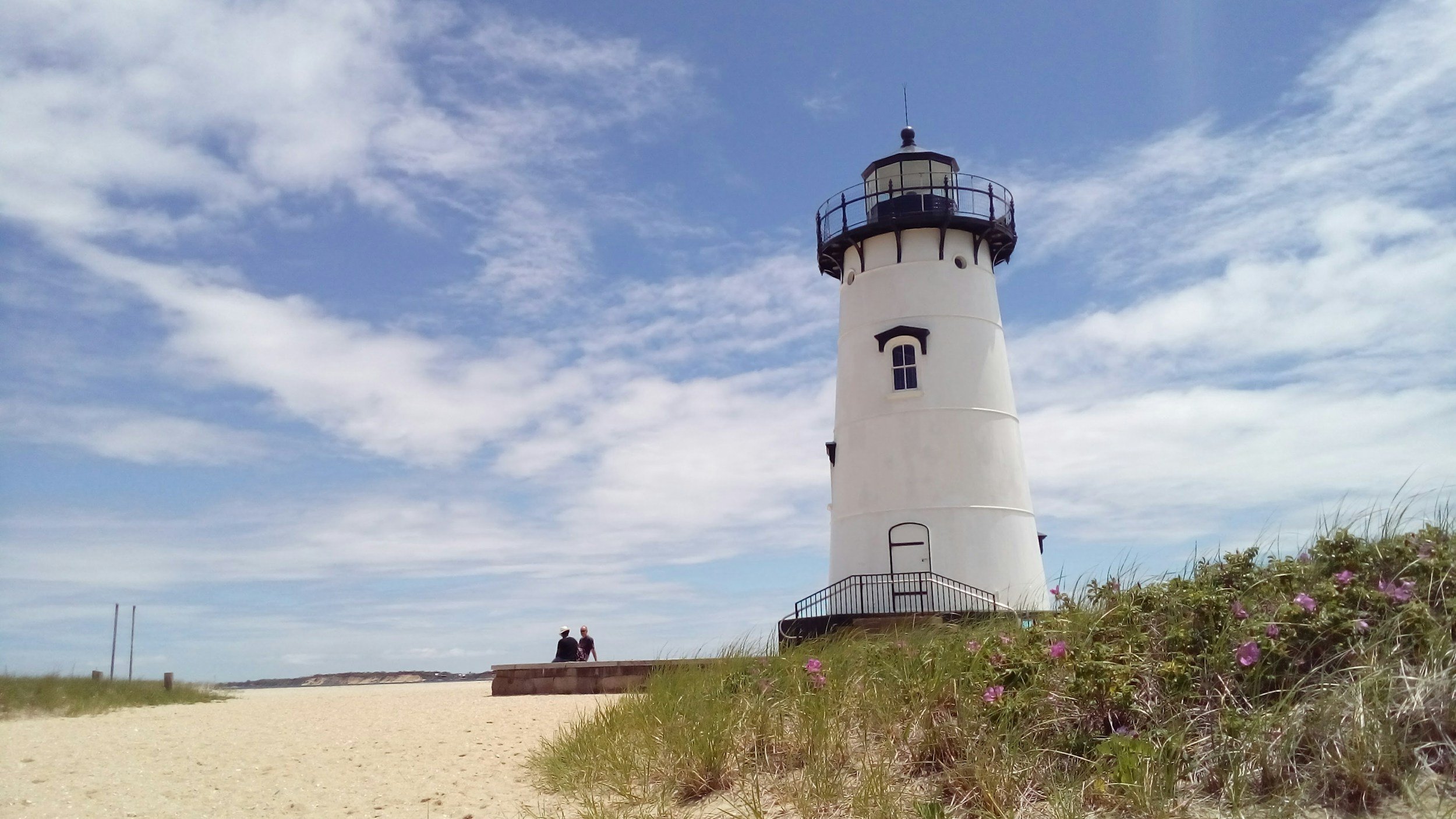 A white lighthouse with black railing and a small balcony at the top, standing on a grassy area with pink flowers, against a partly cloudy blue sky, overlooking a sandy beach.