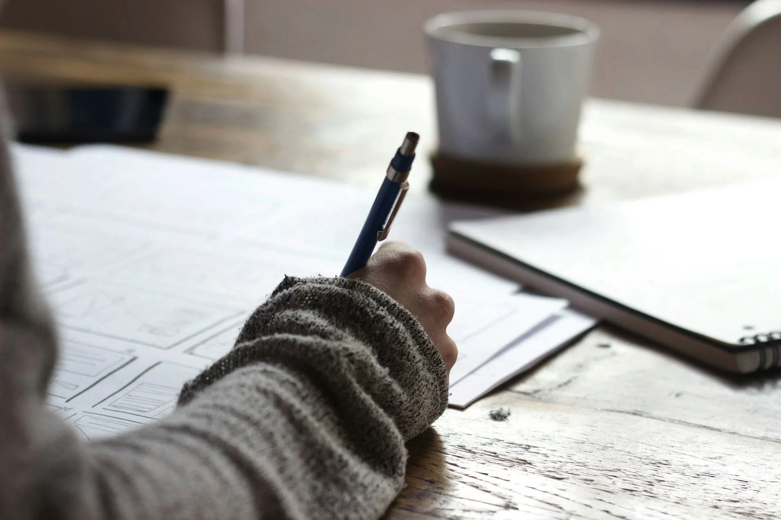 Close-up of a person's hand holding a blue pen, writing on papers with diagrams or charts, on a wooden table with a coffee mug and a notebook in the background.