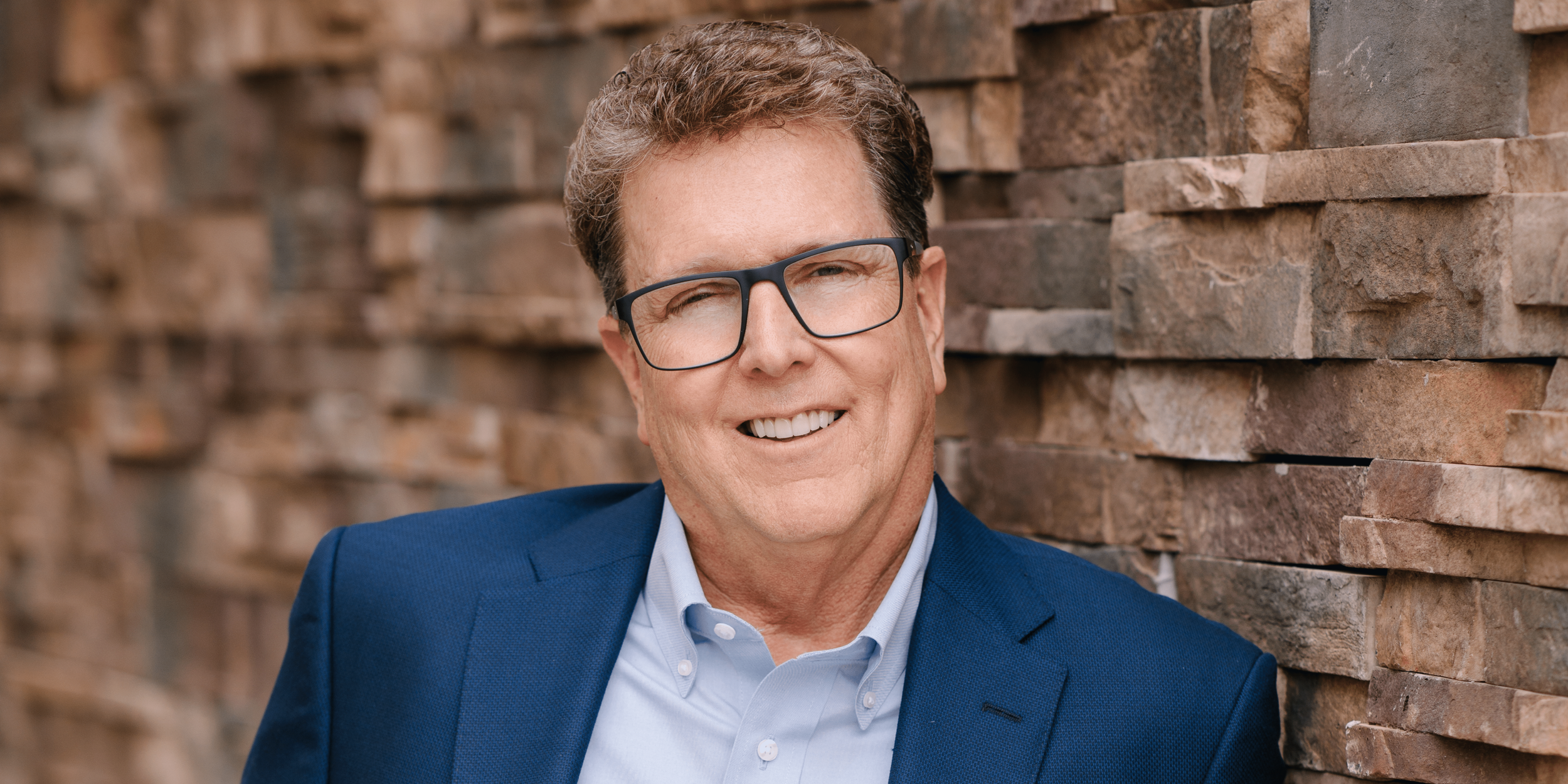Man wearing glasses and a blue suit jacket, smiling, standing against a brick wall.