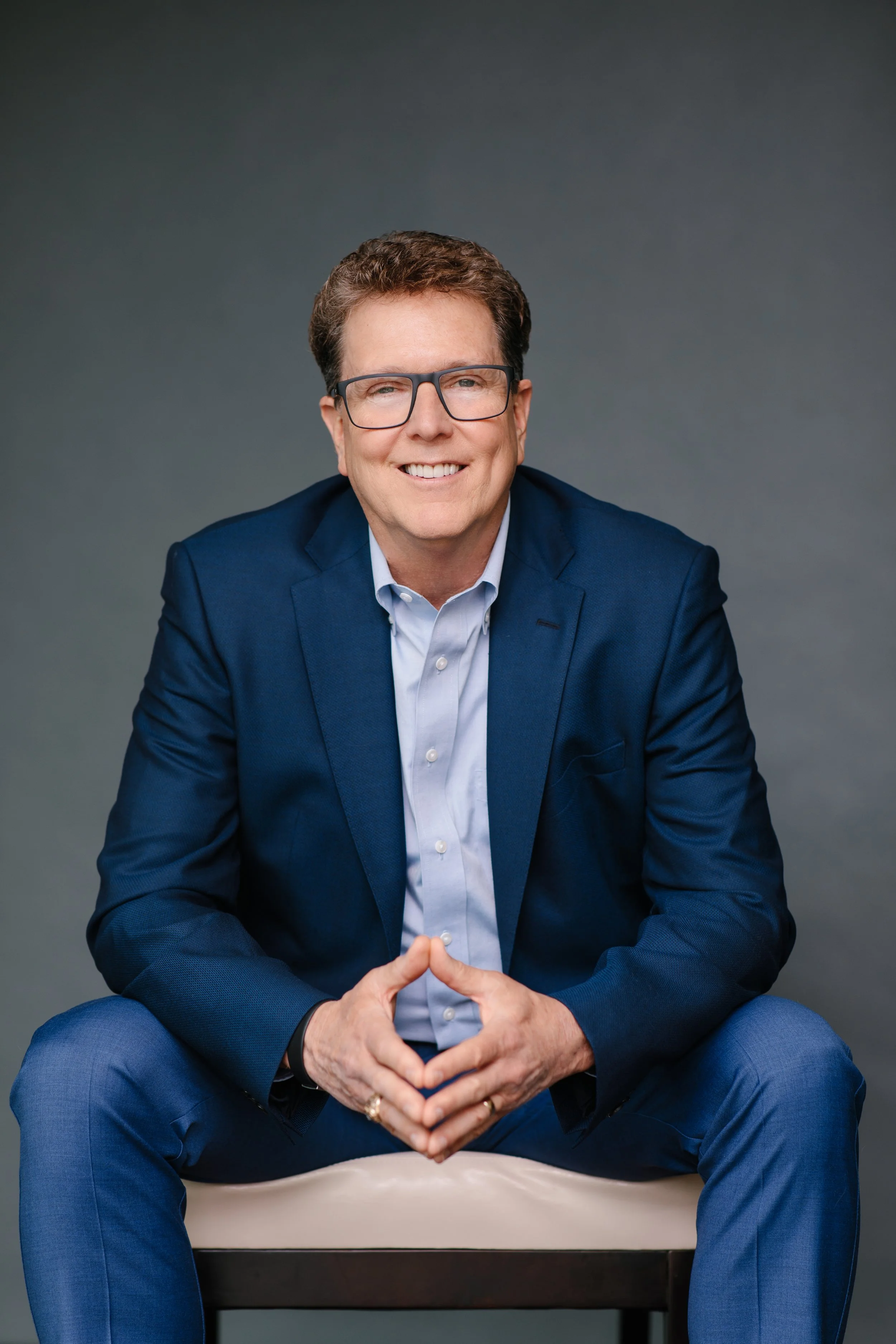 A man in a blue suit sits on a beige chair, smiling at the camera, with a gray background.