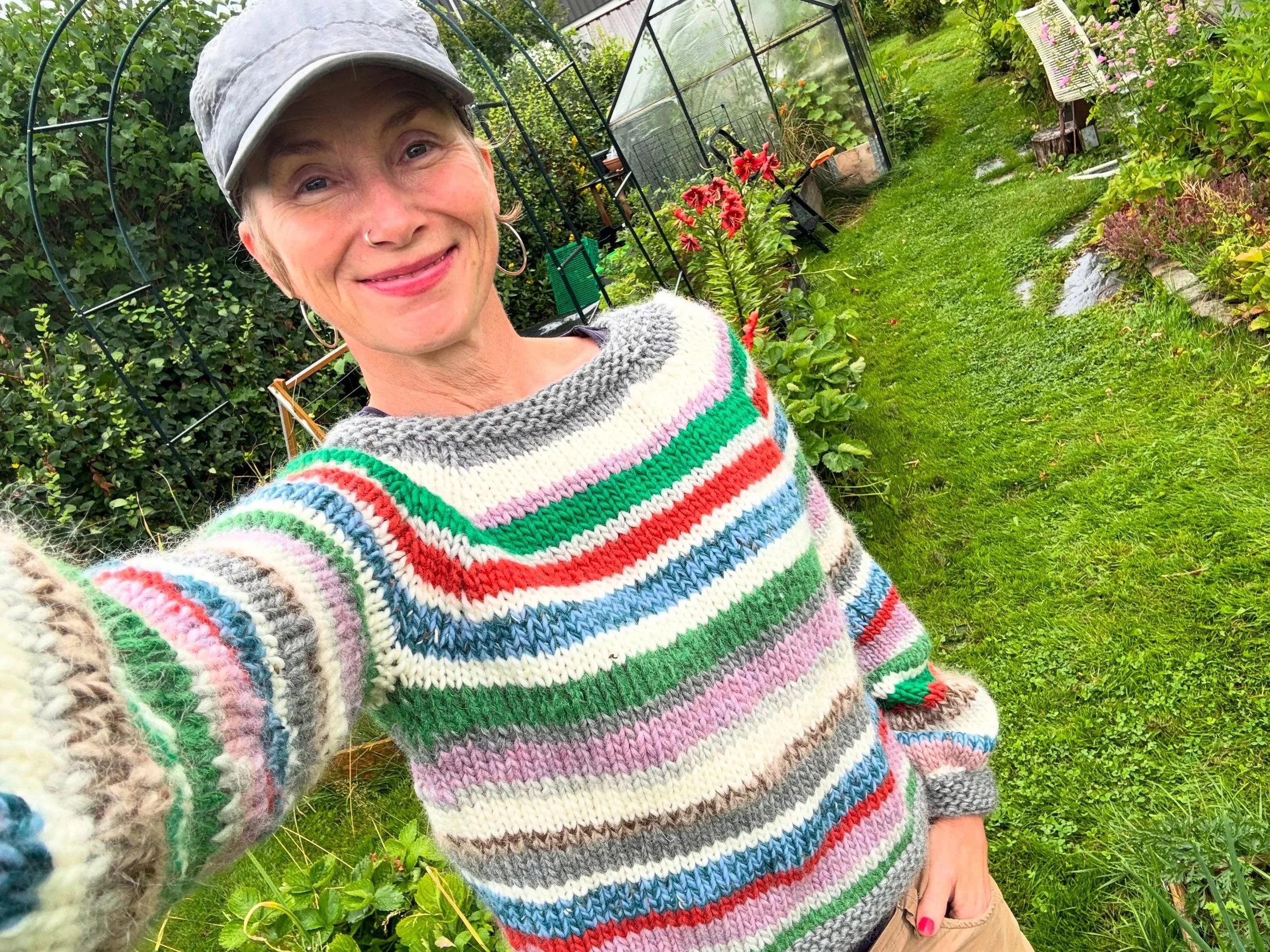 En kvinne tar et selfie i en hage, omgitt av blomster, grønnsaker og et drivhus, iført en stripet, fargerik strikket genser og en grå caps.