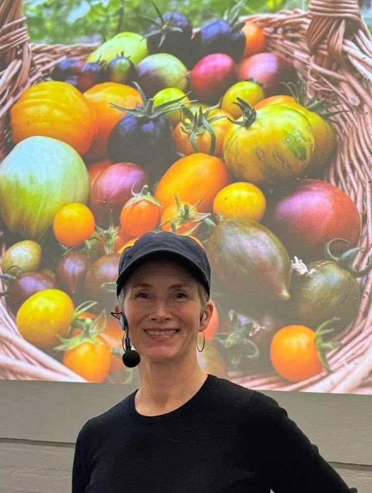 Et bilde av en kvinne med en mikrofon foran en stor skjerm som viser en kurv med forskjellige fargerike tomater.